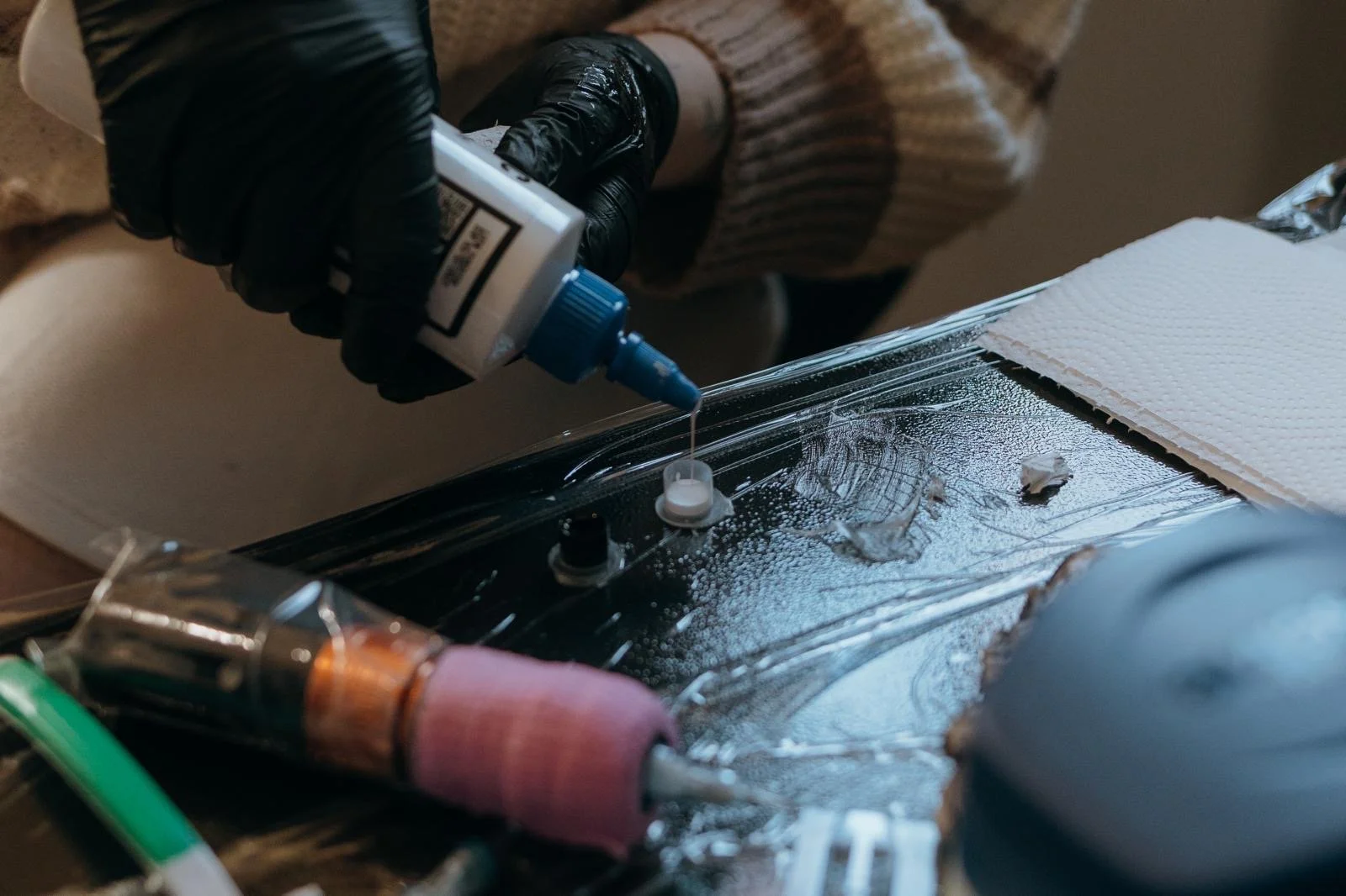 A person wearing black gloves is applying liquid onto a glass surface using a squeeze bottle. The glass is secured with a suction cup and surrounded by various medical or scientific instruments and cloths.