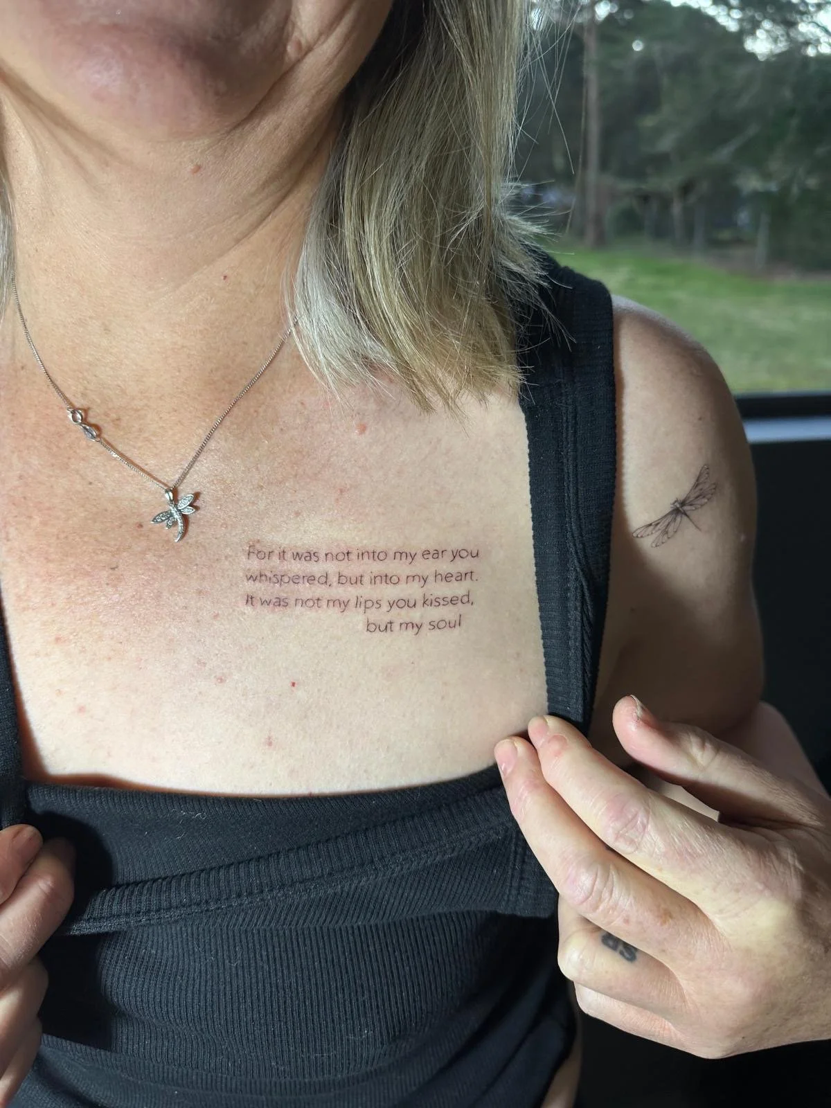 A woman in a black tank top shows a tattooed quote on her chest and another small tattoo of a dragonfly on her shoulder. She wears a necklace with a butterfly pendant.
