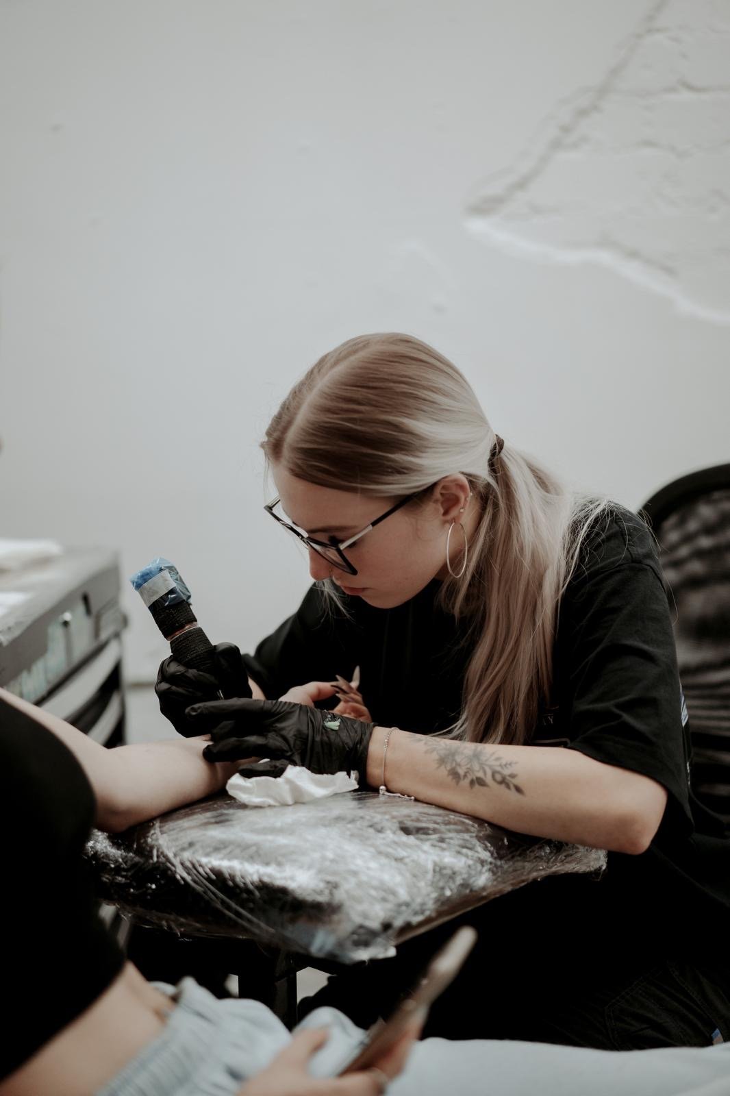 A woman with blonde hair, glasses, and tattoos on her arm is tattooing a person's hand in a tattoo studio.
