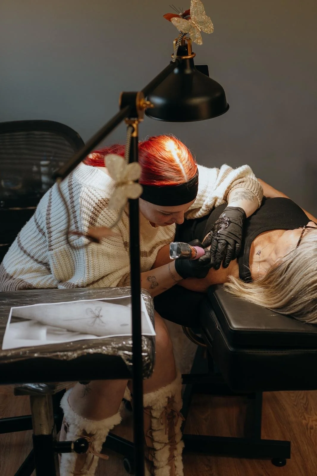 A tattoo artist with red hair, wearing a black headband and black gloves, tattooing an older woman's arm while she is lying on a black tattoo chair.