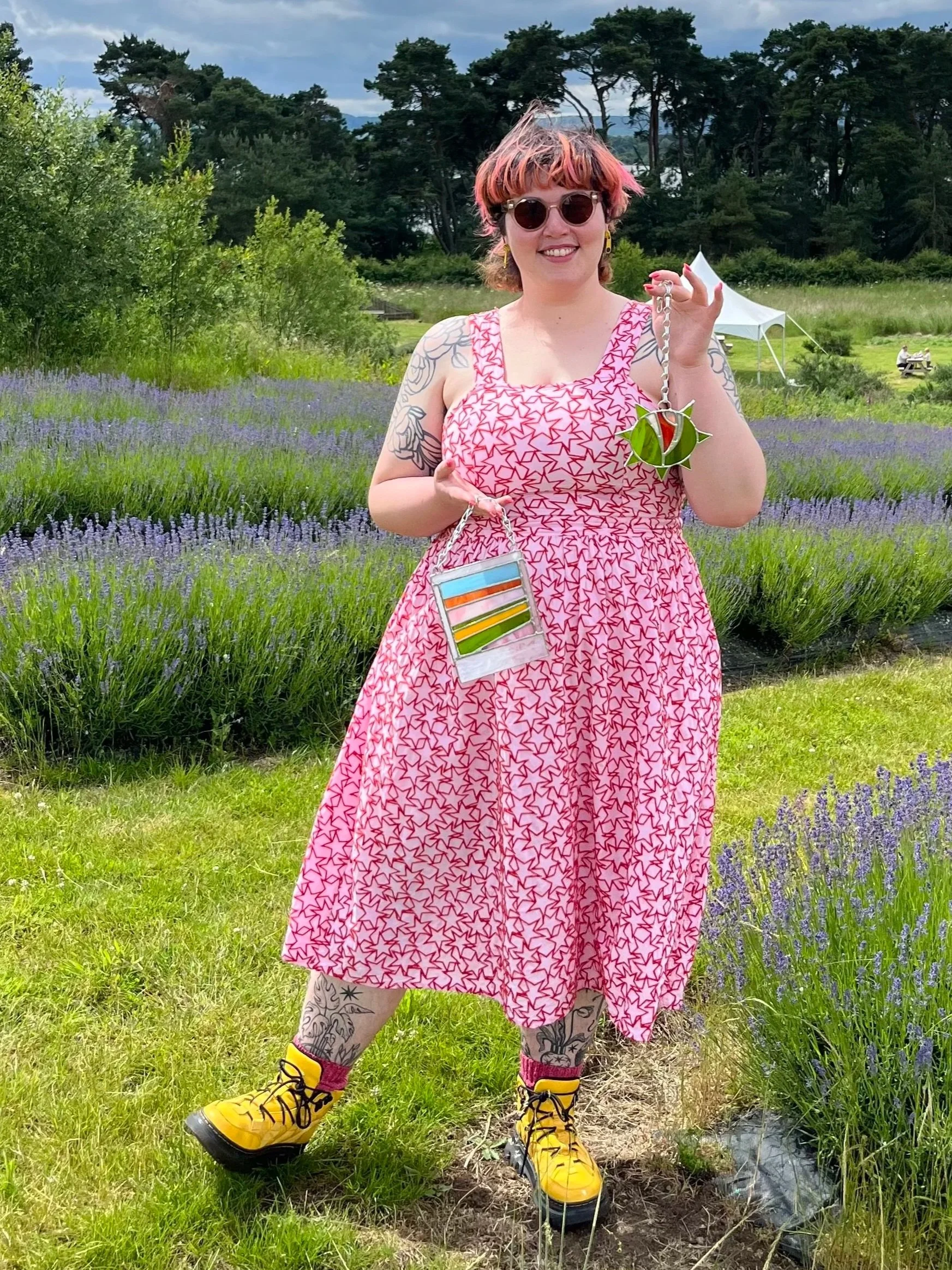 A woman with pink and orange hair, wearing round sunglasses, a pink dress with star print, yellow hiking boots, and tattoos on her arms and leg, stands outdoors on a grassy area with purple flowers in a natural park setting with trees and cloudy sky in the background.