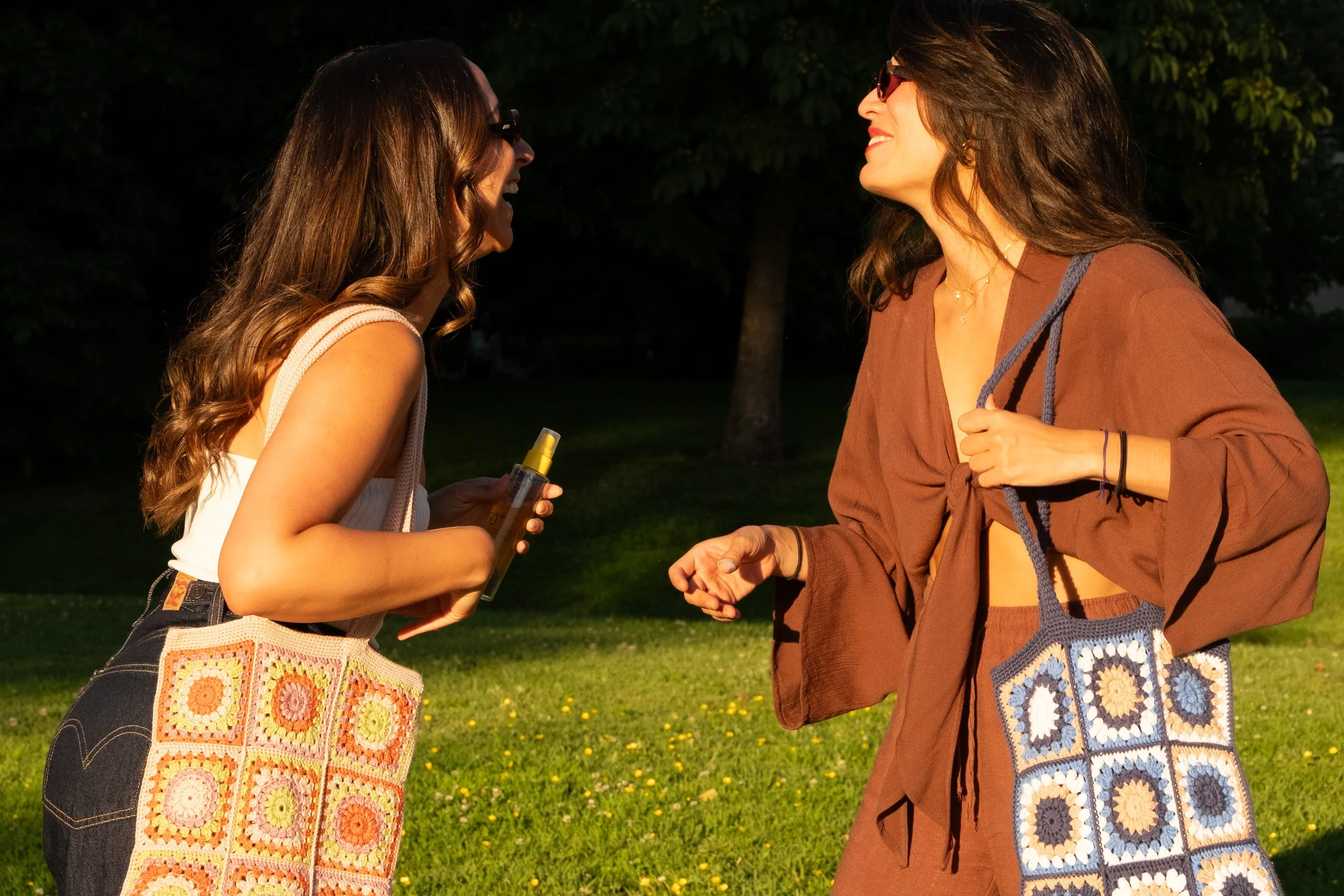 Deux femmes souriantes discutent dans un parc en soirée, l'une portant un sac crocheté coloré et l'autre avec un ensemble marron et un sac en crochet bleu artisanal
