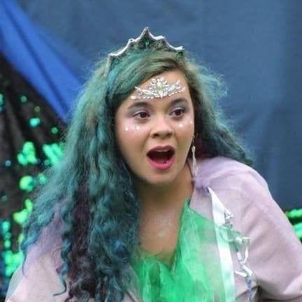 Rachael - a woman  with long, curly blue hair, wearing a tiara and a pastel-colored costume, looking surprised or shocked.