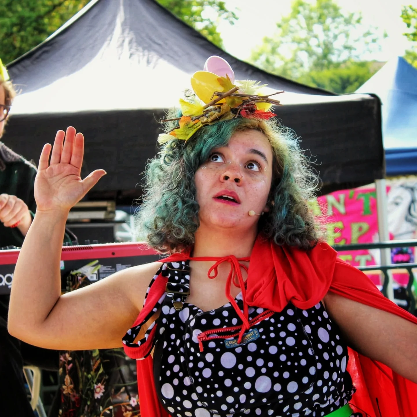 Rachael - a woman with curly, blue hair wearing a headpiece made of leaves, twigs, and pastel-colored eggs, standing outdoors under trees, raising her right hand with her palm facing out, and wearing polka dot dungarees with a red cape.