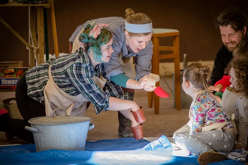 Rachael and her colleague performing in a show - crouched down and engaging with a group of children and an adult. Rachael is making a puppet of a dog using flower pots on her hands as paws, whilst her colleague makes two brown pots and a spade face