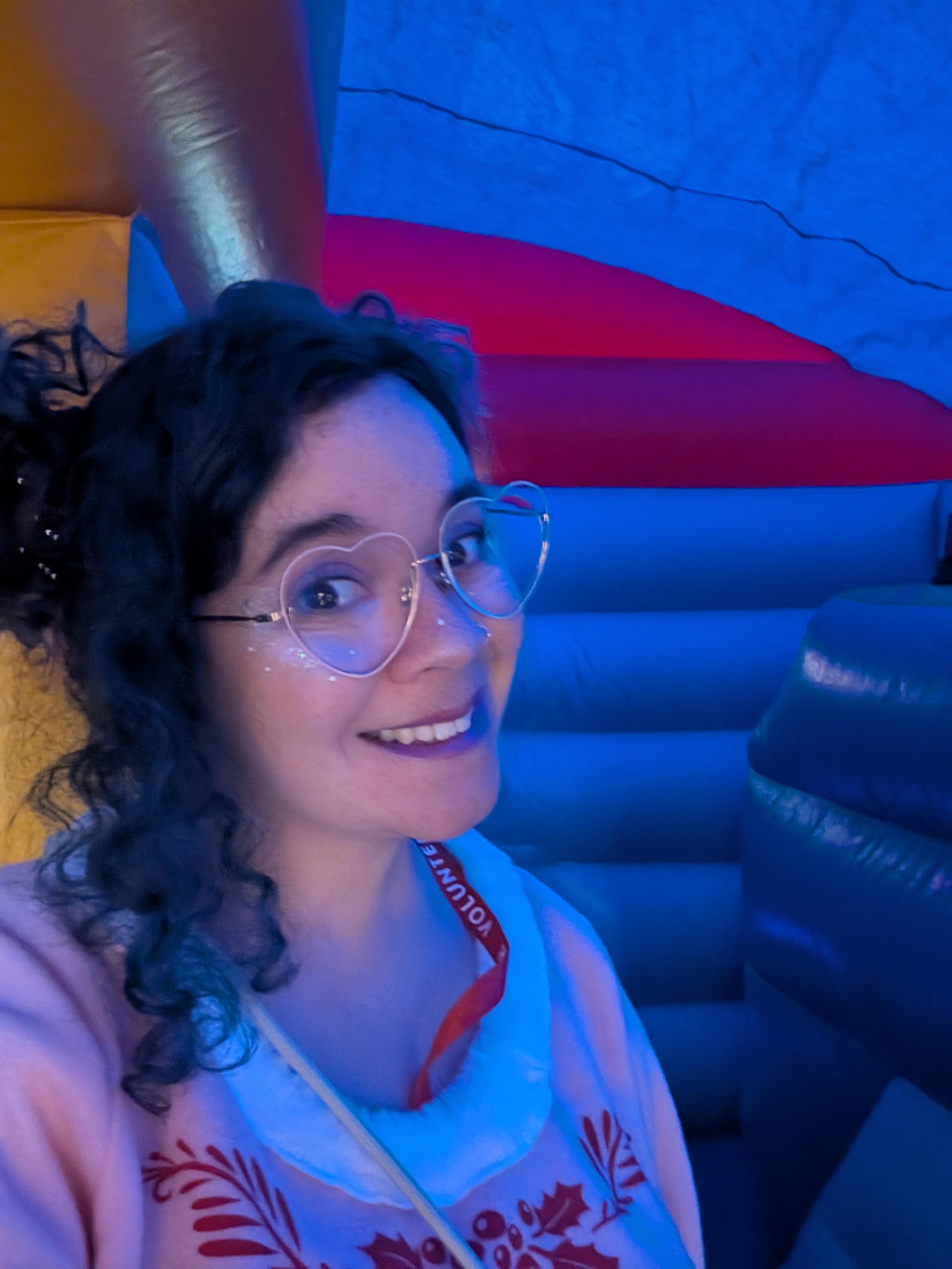 Rachael - a woman with curly  hair and glasses smiling, wearing a pastel pink elf costume, with a red lanyard around her neck, in an inflatable setting with blue lighting and colorful background.