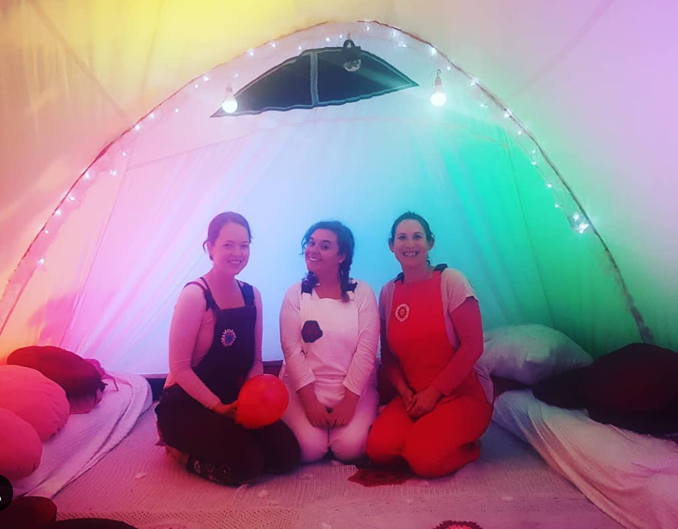Three women sitting cross-legged inside a colorful, rainbow-lit tent with pillows and blankets, smiling at the camera.