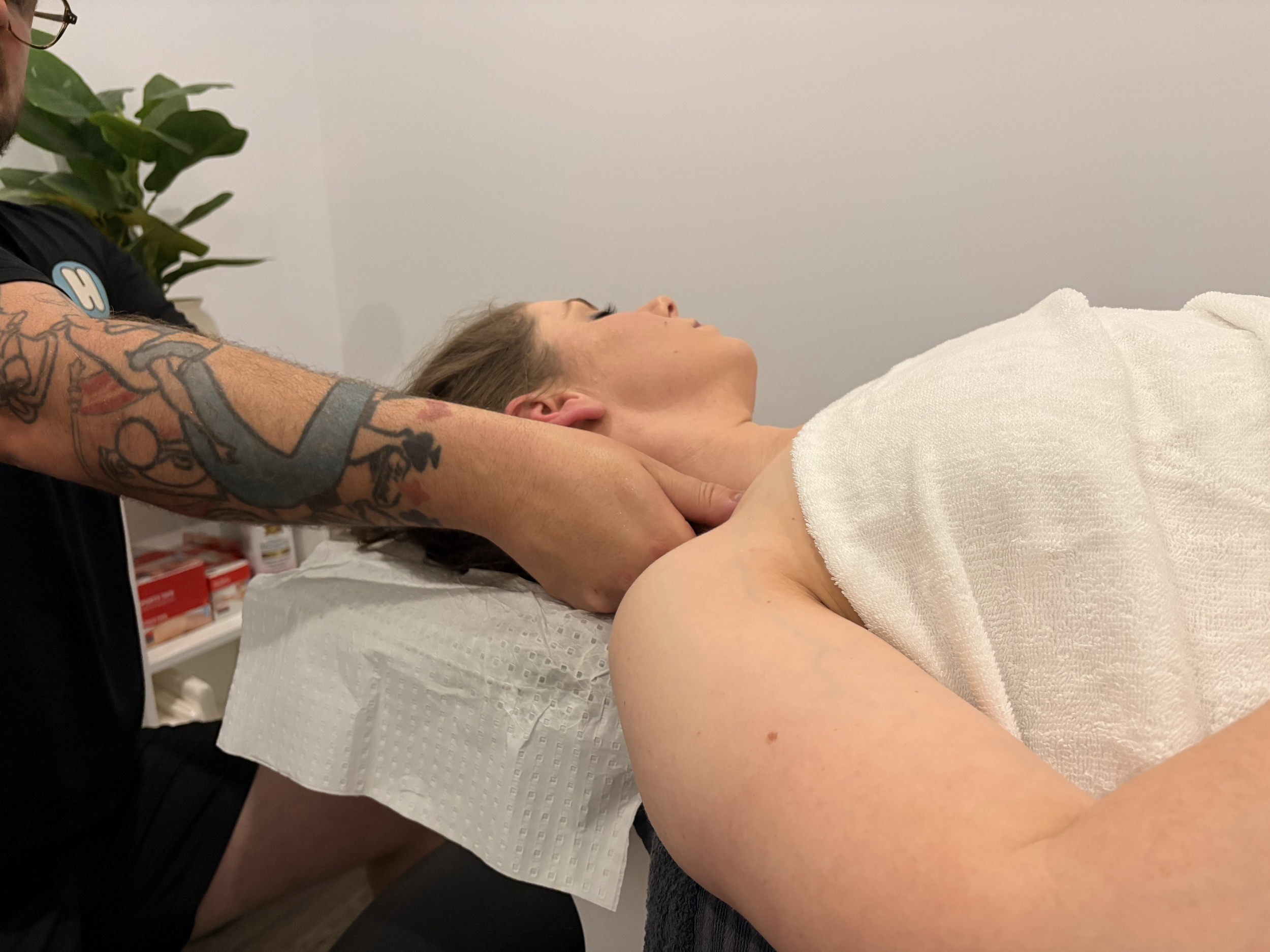 A woman receiving a massage from a massage therapist, lying on a massage table with a white towel covering her body, in a clinical massage room.