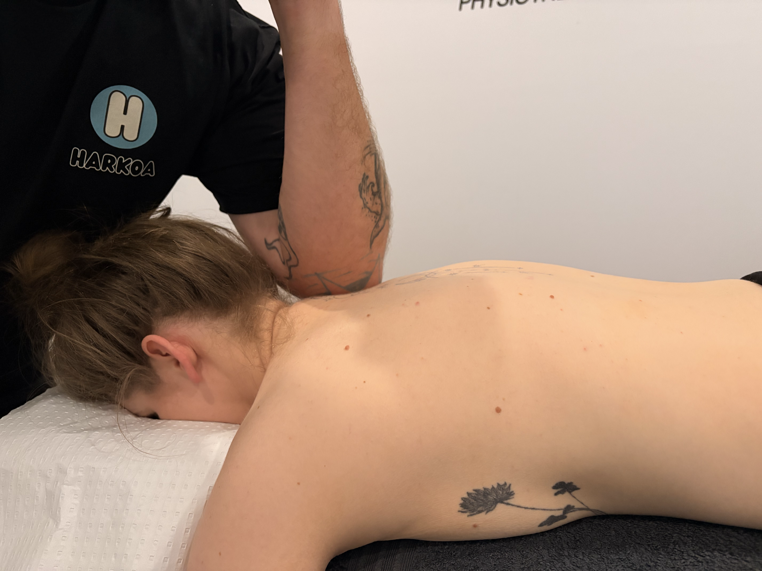 A person lying face down on a white massage table receiving a massage from a therapist. The person has multiple small moles and a tattoo of floral design on their lower back. The therapist is wearing a black shirt with a logo that says 'HARKOA' and has a tattoo of a lion on their arm. The room has a plain white wall in the background.