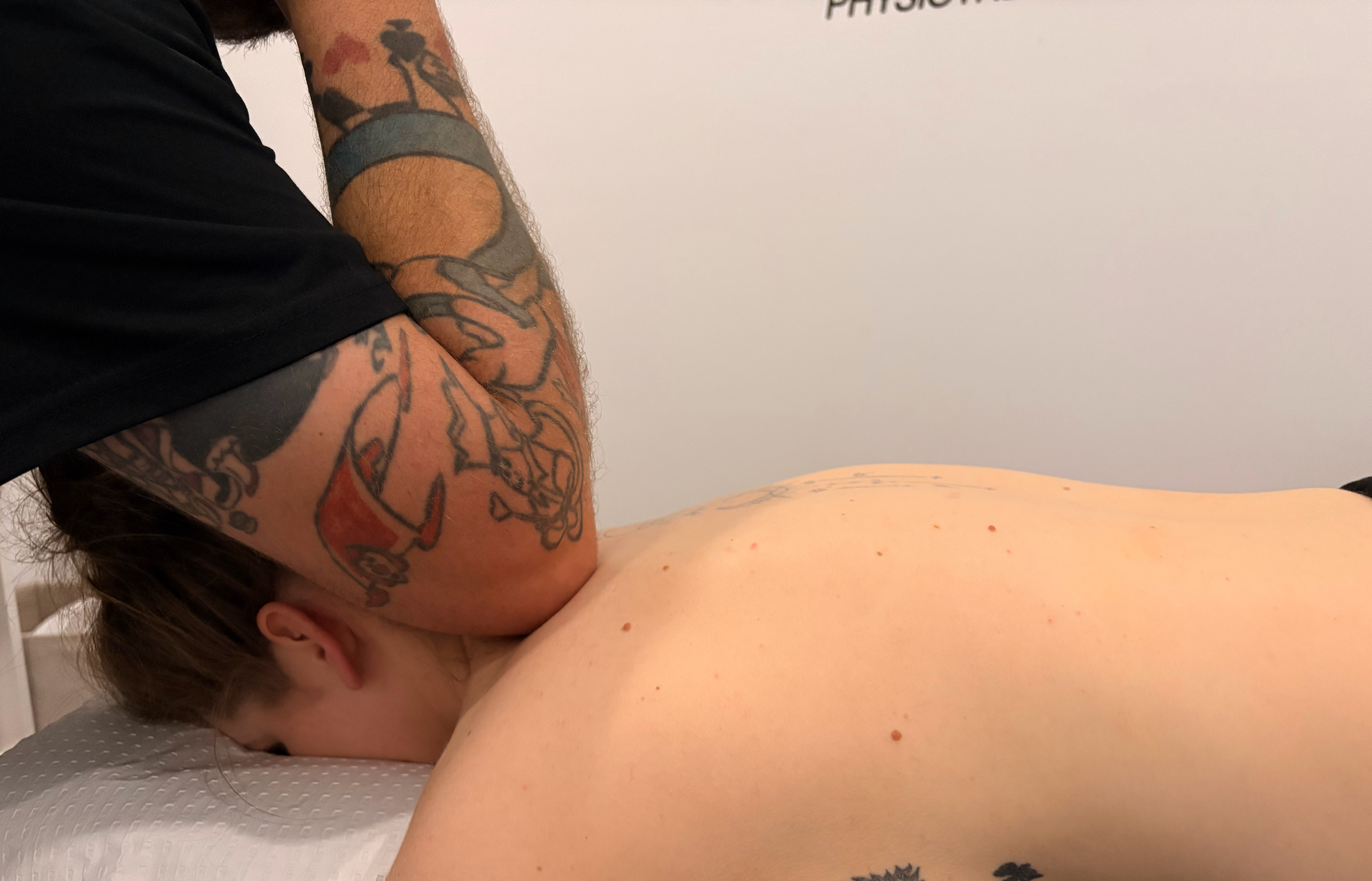 A person receiving a massage or therapy, with visible tattoos on their arm, lying face down on a massage table with a white textured cover, in a room with a plain white wall.