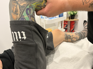 A person lying on a medical examination table with tattoos on their arm and torso, wearing black shorts with white lettering.