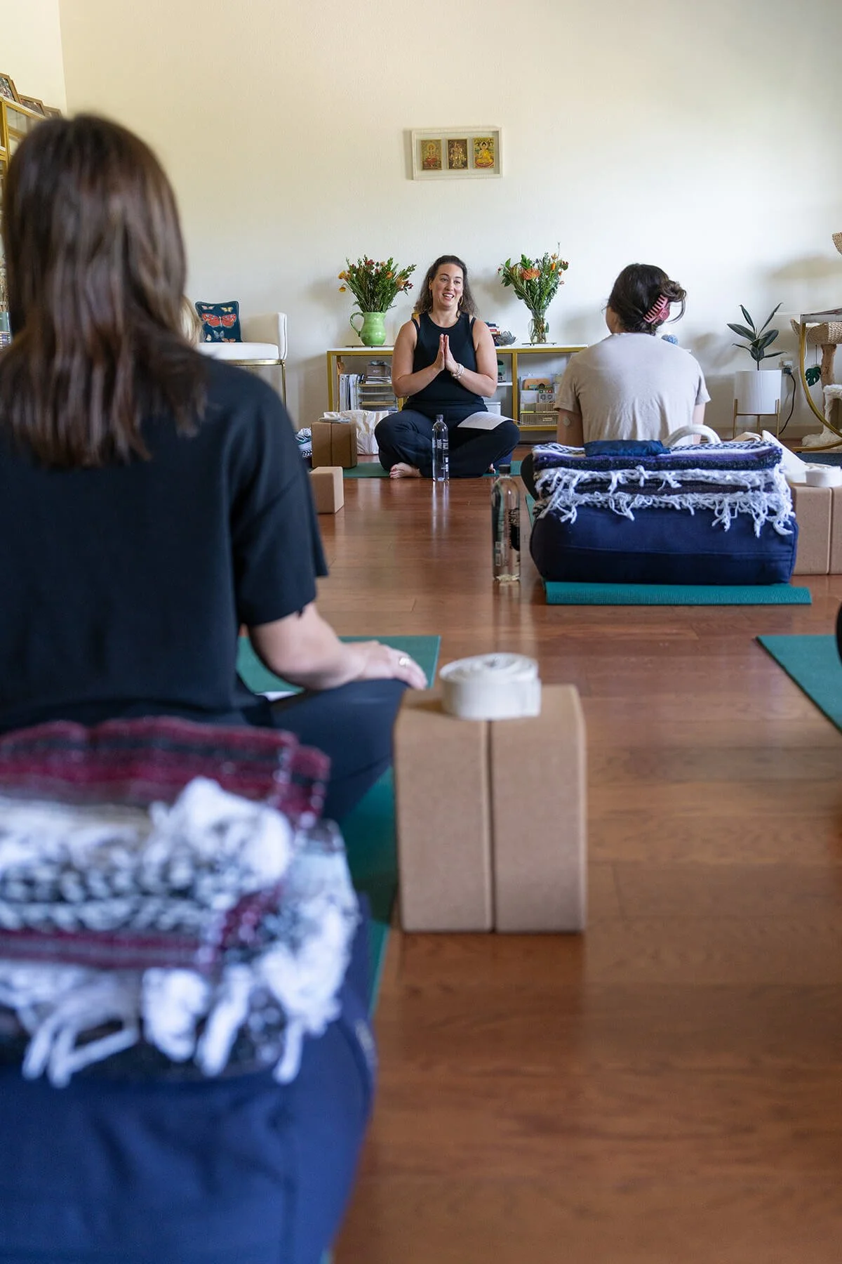 Women sitting on yoga mats in a circle in a well-lit living room with flowers and artwork, participating in a group meditation or yoga session.