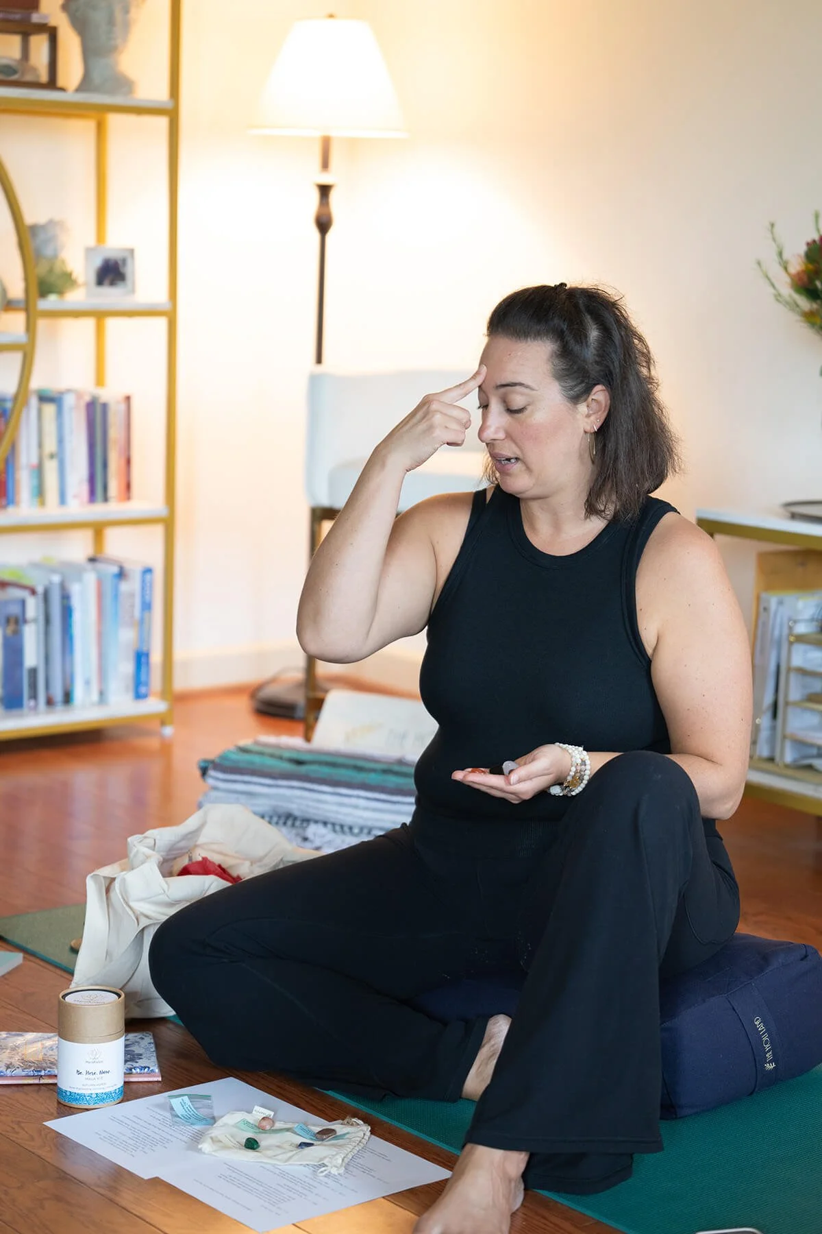 Woman sitting on a cushion indoors, holding a stone in her hand, with various items and papers on the floor, a bookcase, a lamp, and a plant in the background.