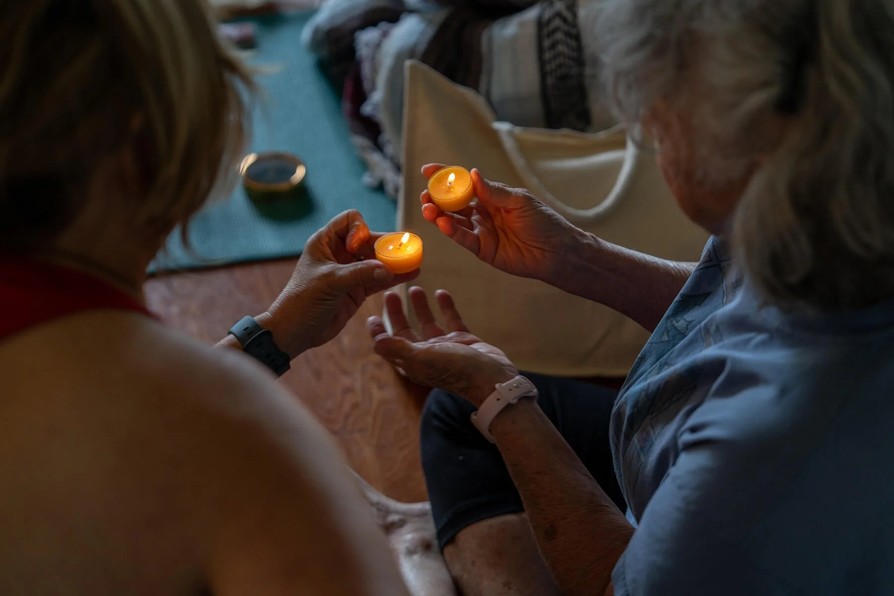 Two elderly women are holding small lit candles, and another woman is sitting nearby. The scene appears to be in a cozy room with a table, a blue mat, and a bowl in the background.
