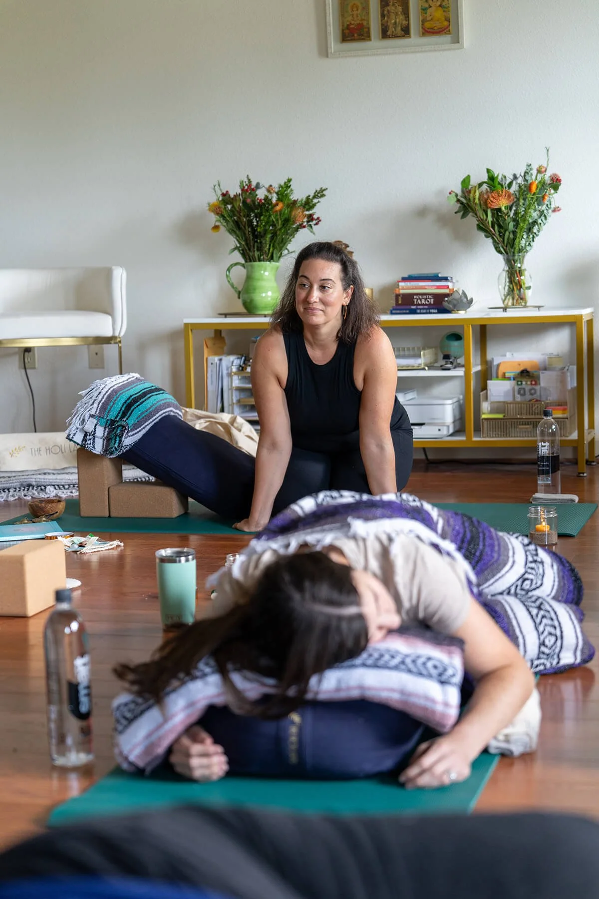 Woman leading a yoga or meditation class with one person lying on a yoga mat in a bright room decorated with flowers and spiritual artwork.