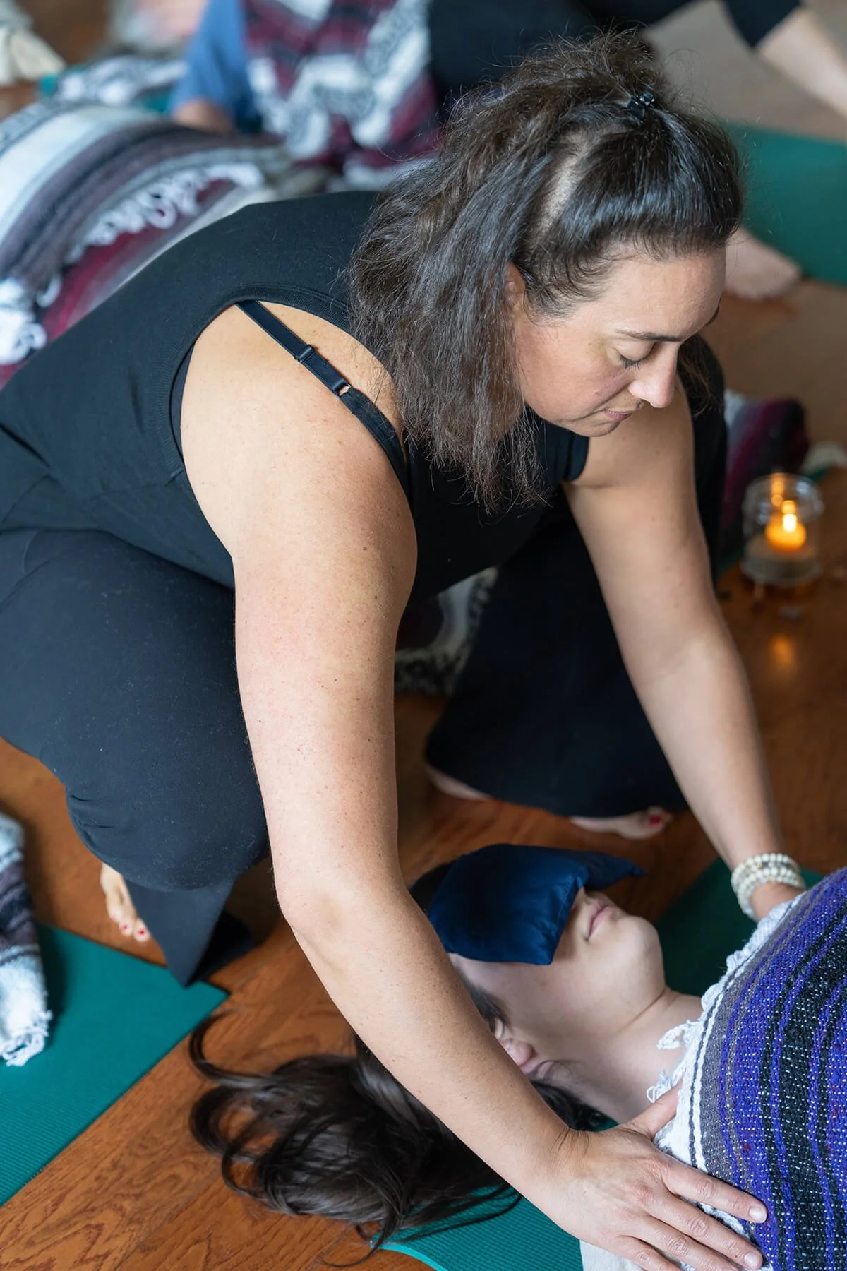 A woman with dark, curly hair performing a yoga pose on a young girl lying on a yoga mat. The girl has long dark hair, a blue eye mask, and is relaxing while the woman supports her with her hands. The setting is indoors with wooden flooring and a candle in the background.