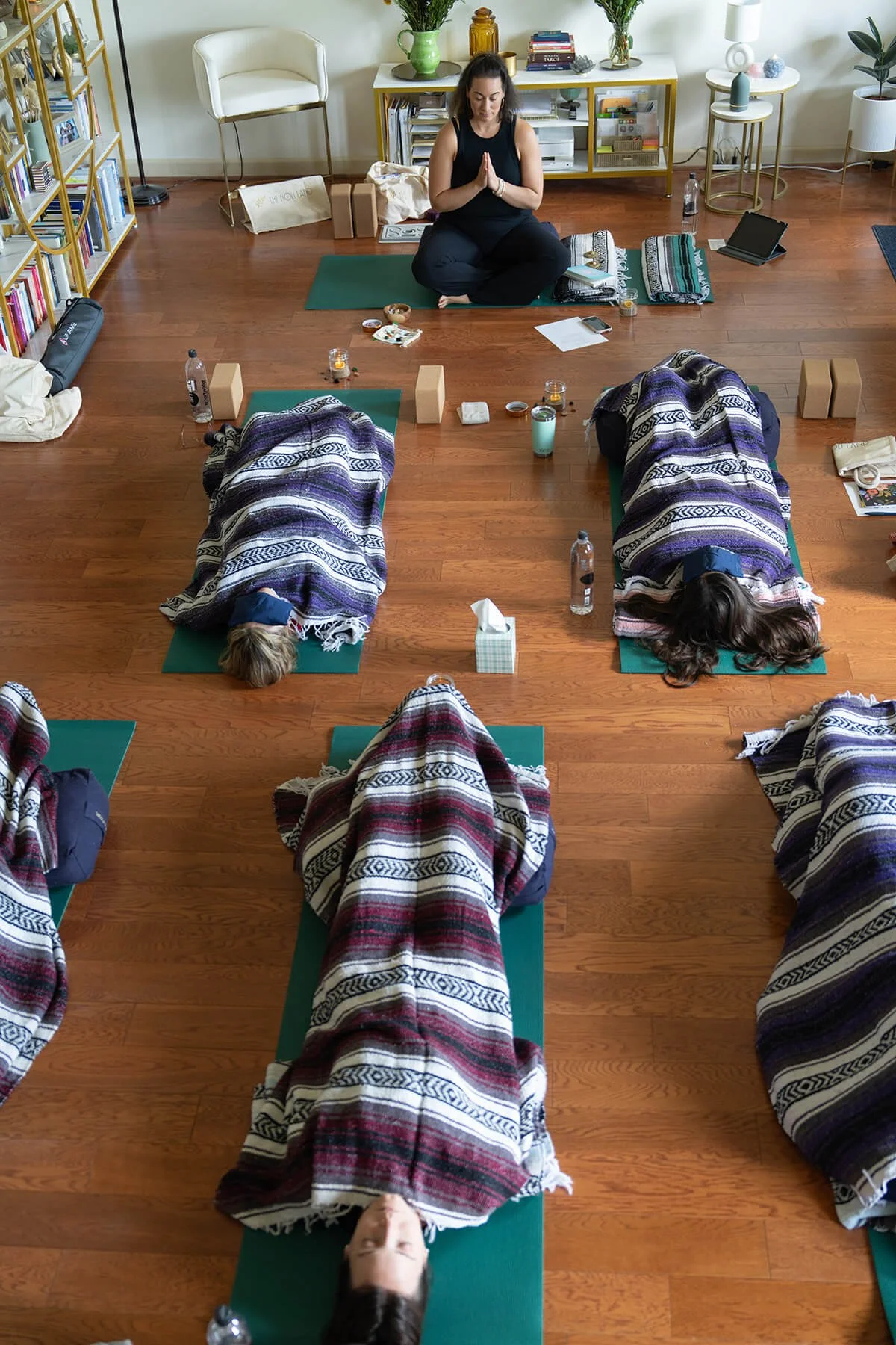 A group yoga or meditation class in a spacious room with wooden floors, featuring four individuals lying on green mats wrapped in blankets, while a woman in a black top sits cross-legged on a mat with hands pressed together in a prayer position. The room is decorated with plants, books, and various decorations.