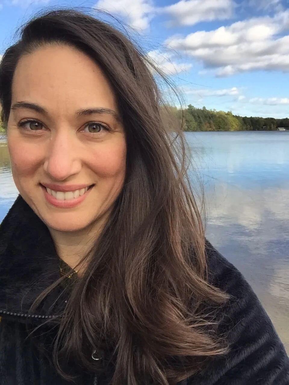 A woman with long brown hair taking a selfie near a body of water on a partly cloudy day.