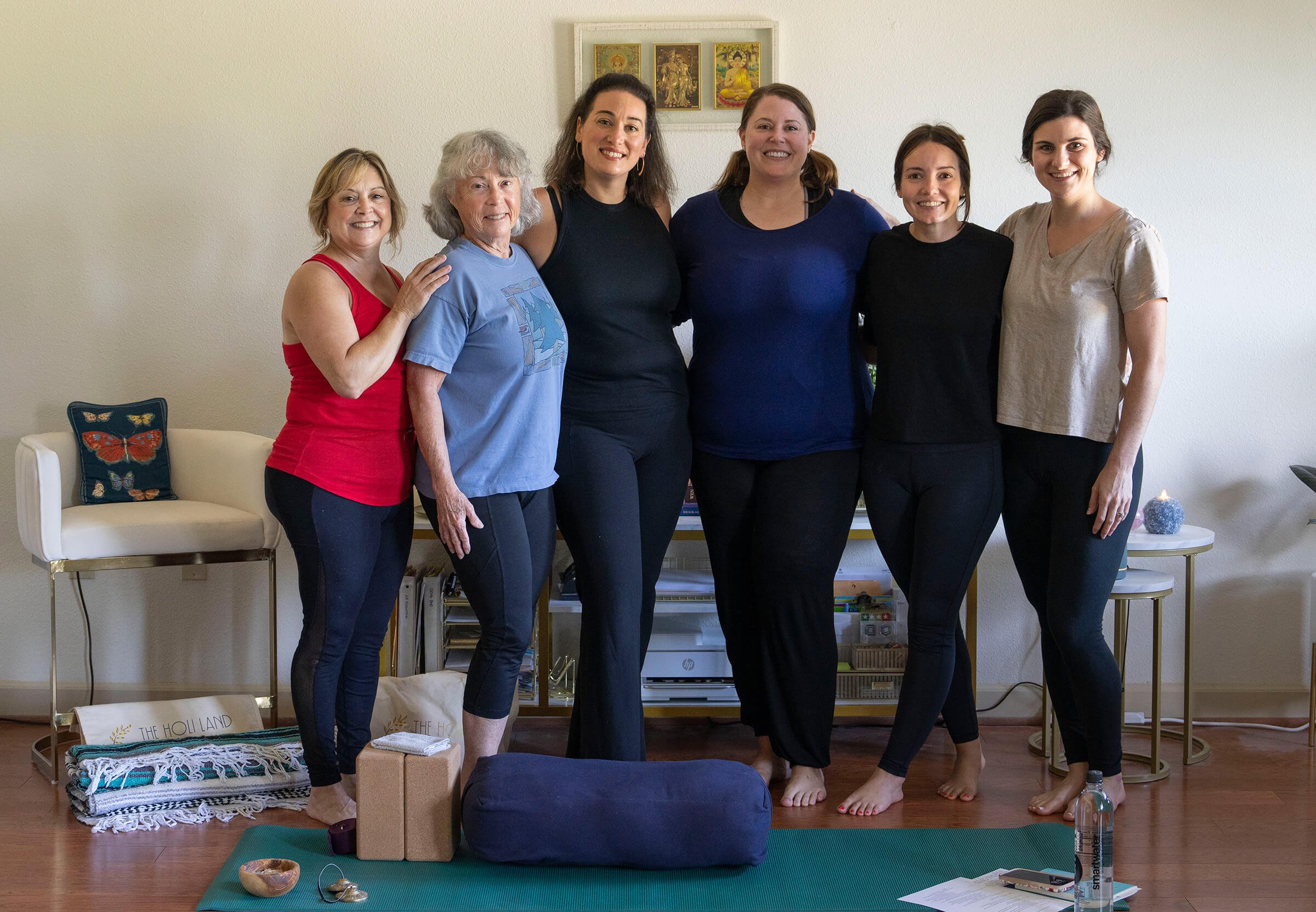 Six women standing together in a yoga studio, smiling at the camera during a yoga or meditation class with mats, blocks, and a water bottle in the foreground.
