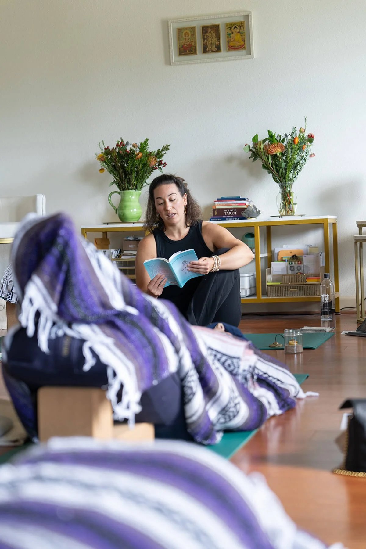 A woman sitting on the floor reading a book in a room with flowers, books, and a water bottle.