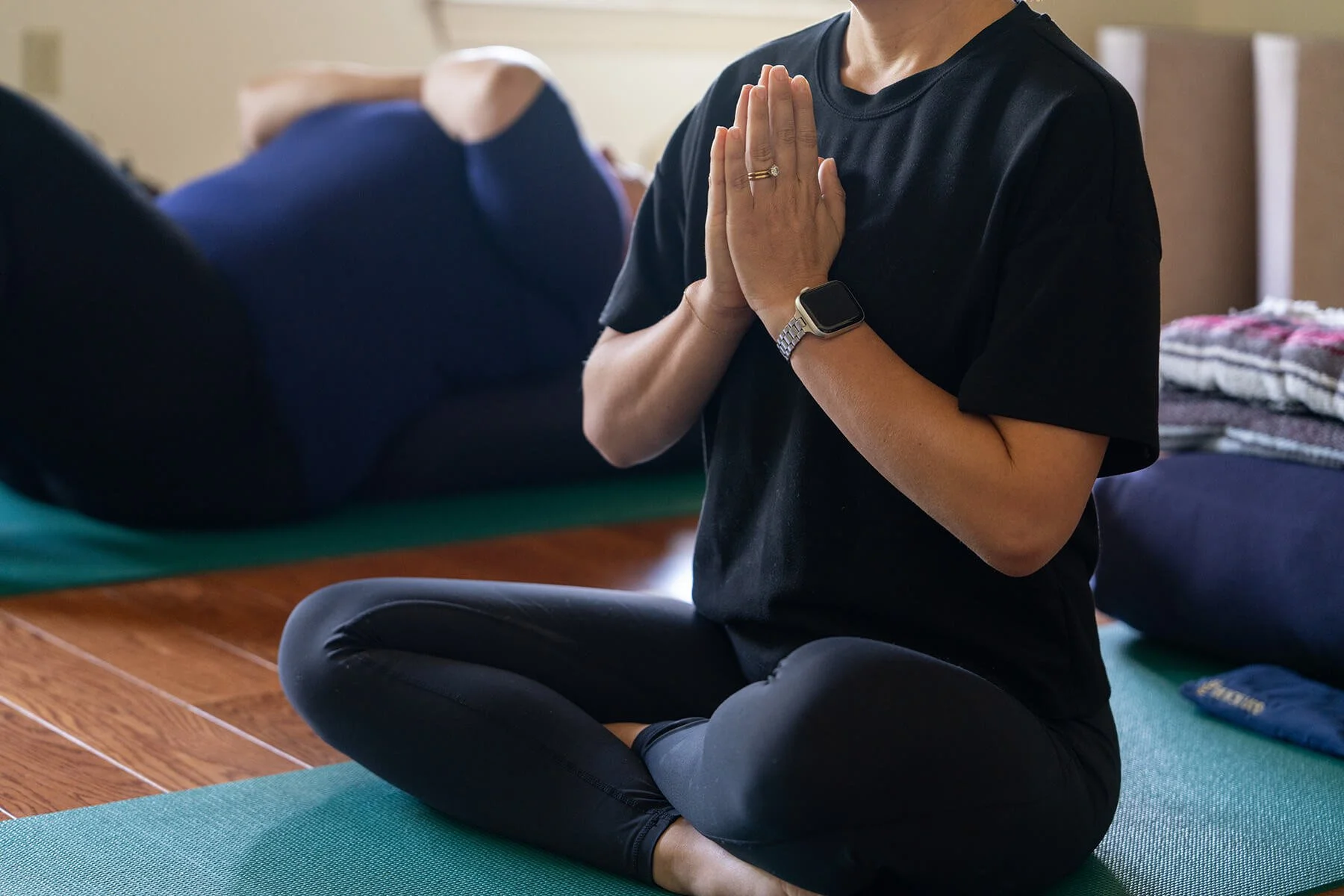 A person practicing yoga indoors in a kneeling meditation pose with hands in prayer position, wearing a black t-shirt, wristwatch, and a ring.