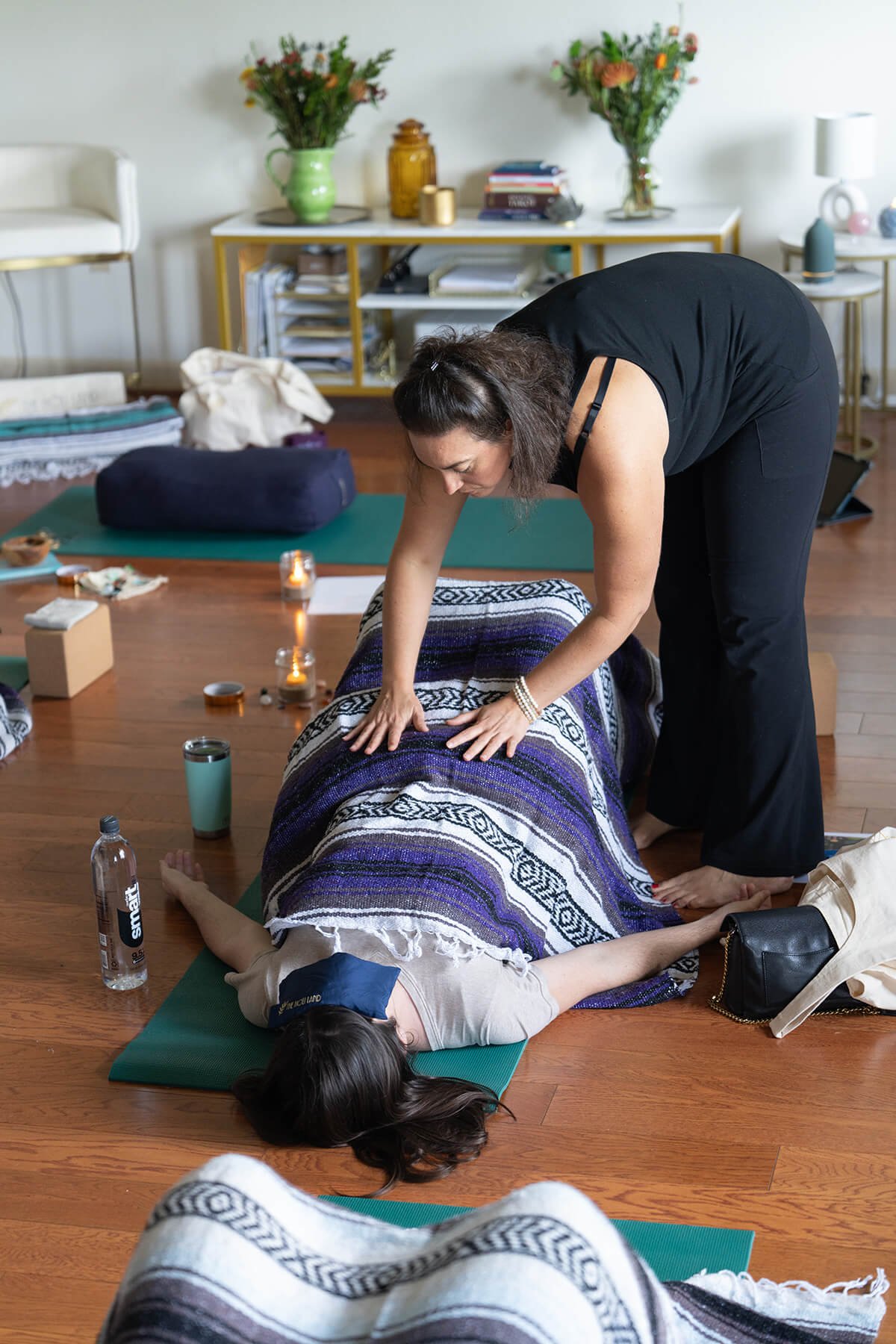 A woman is giving a massage to a person lying face up on a yoga mat in a room decorated with flowers and candles.