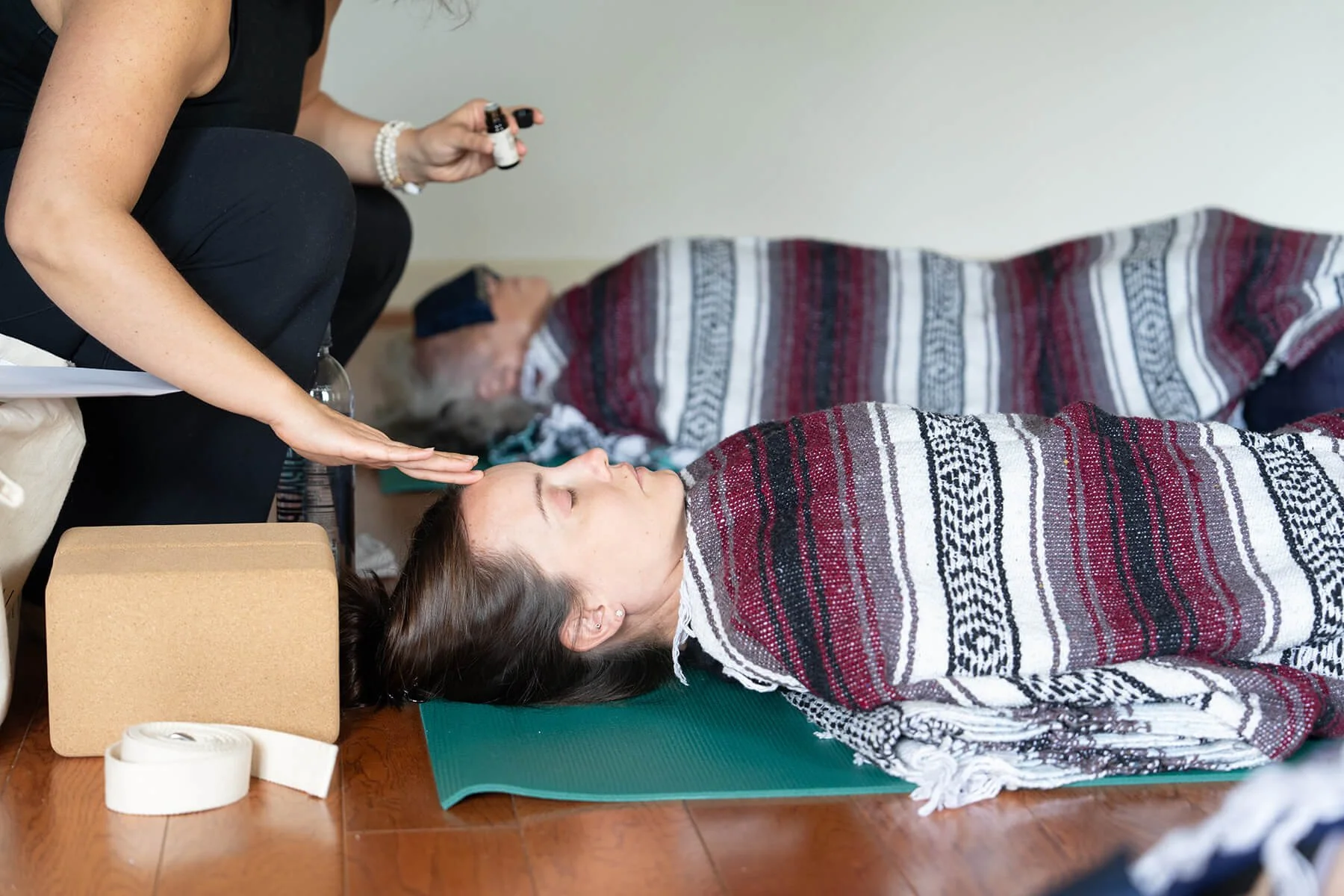A woman lying on her back on a green yoga mat during a sound healing session with singing bowls in the background. A practitioner is seated beside her, holding a small bottle, possibly of essential oil, and gently placing her hand on the woman's forehead. The woman has her eyes closed and appears relaxed. The setting is a calm indoor space with a blanket draped over the woman.
