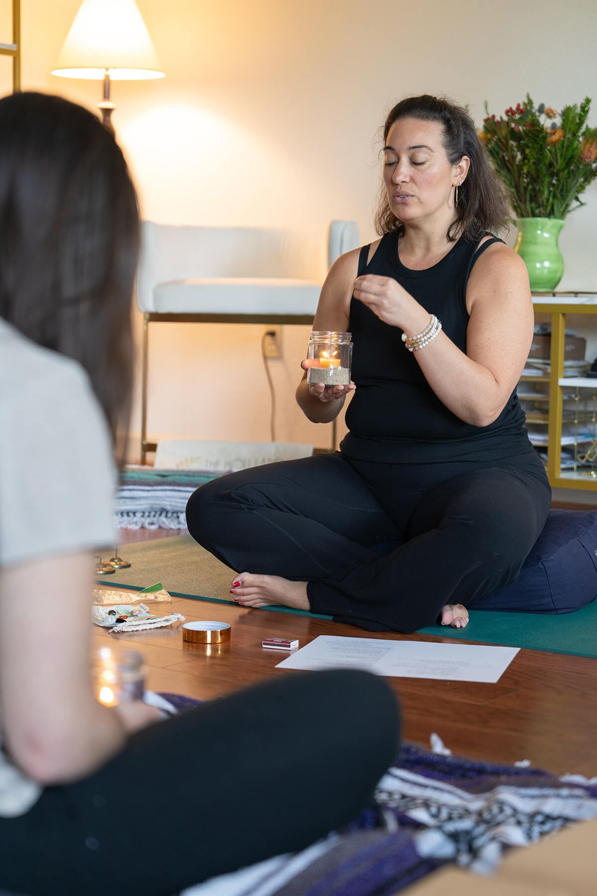 A woman sitting cross-legged on a yoga mat, holding a candle jar, engaging with another person whose back is to the camera. The setting appears to be a cozy living room with a lamp, a plant in a green pot, and various items on a table.