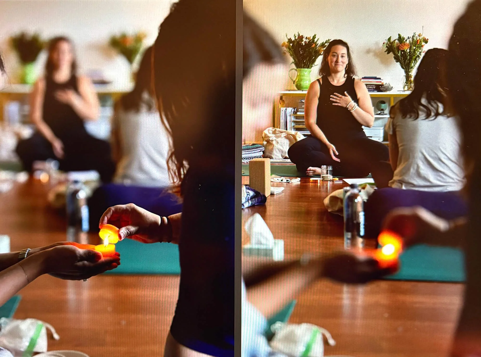 Two women sitting cross-legged on the floor, participating in a meditation or mindfulness session. One woman has dark hair, is wearing a black tank top, and has her hand over her chest. The other woman, with dark hair, is turning away from the camera. In the foreground, a person's hand holds a small lit candle, and there are flowers and books in the background.