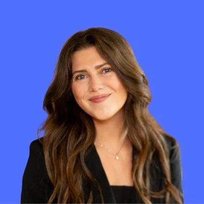 A woman with long, wavy brown hair wearing a black blazer and a necklace, smiling against a blue background.