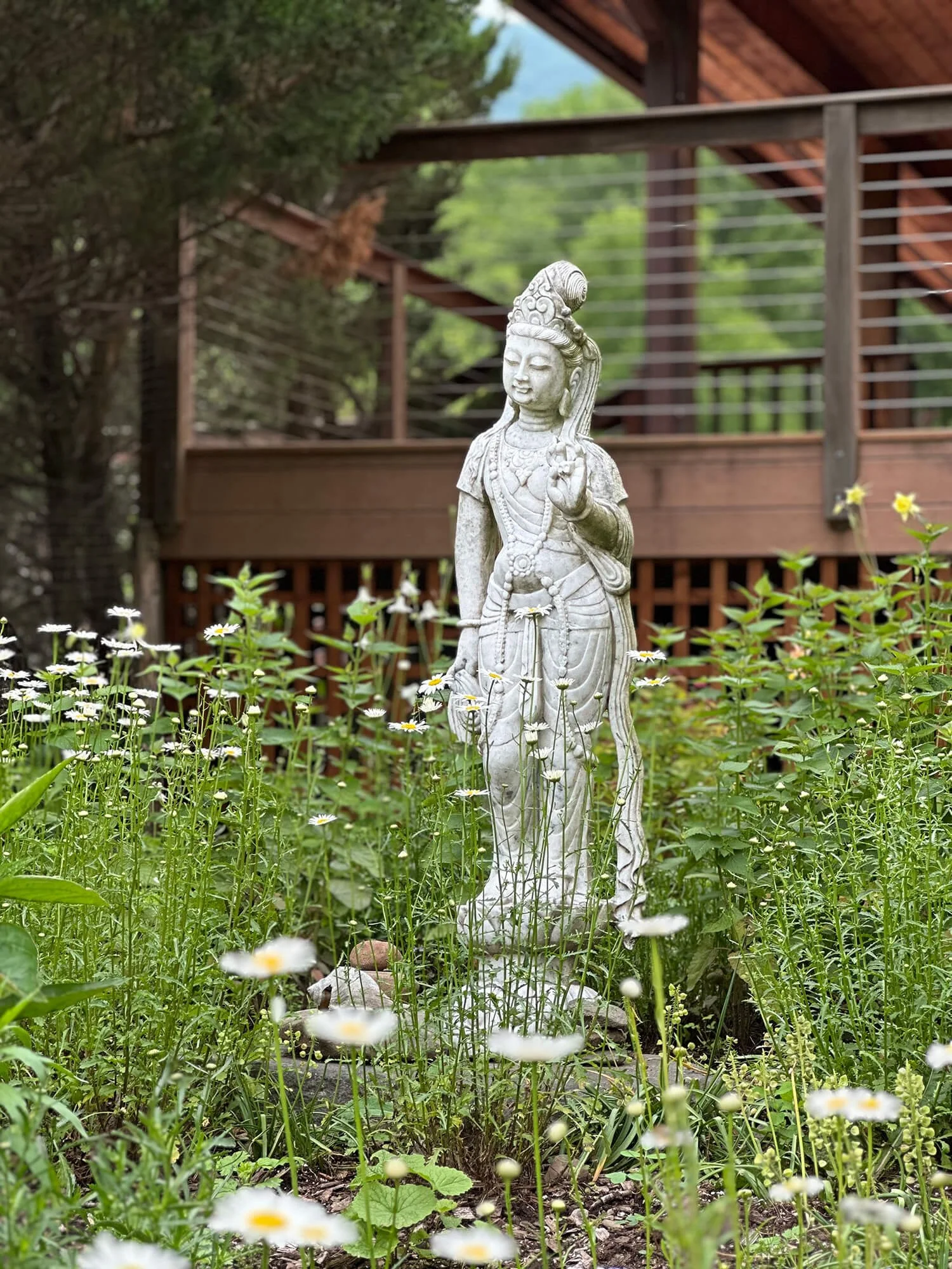 A white garden statue of a woman with intricate jewelry, standing amidst small white flowers, with a wooden balcony and trees in the background.