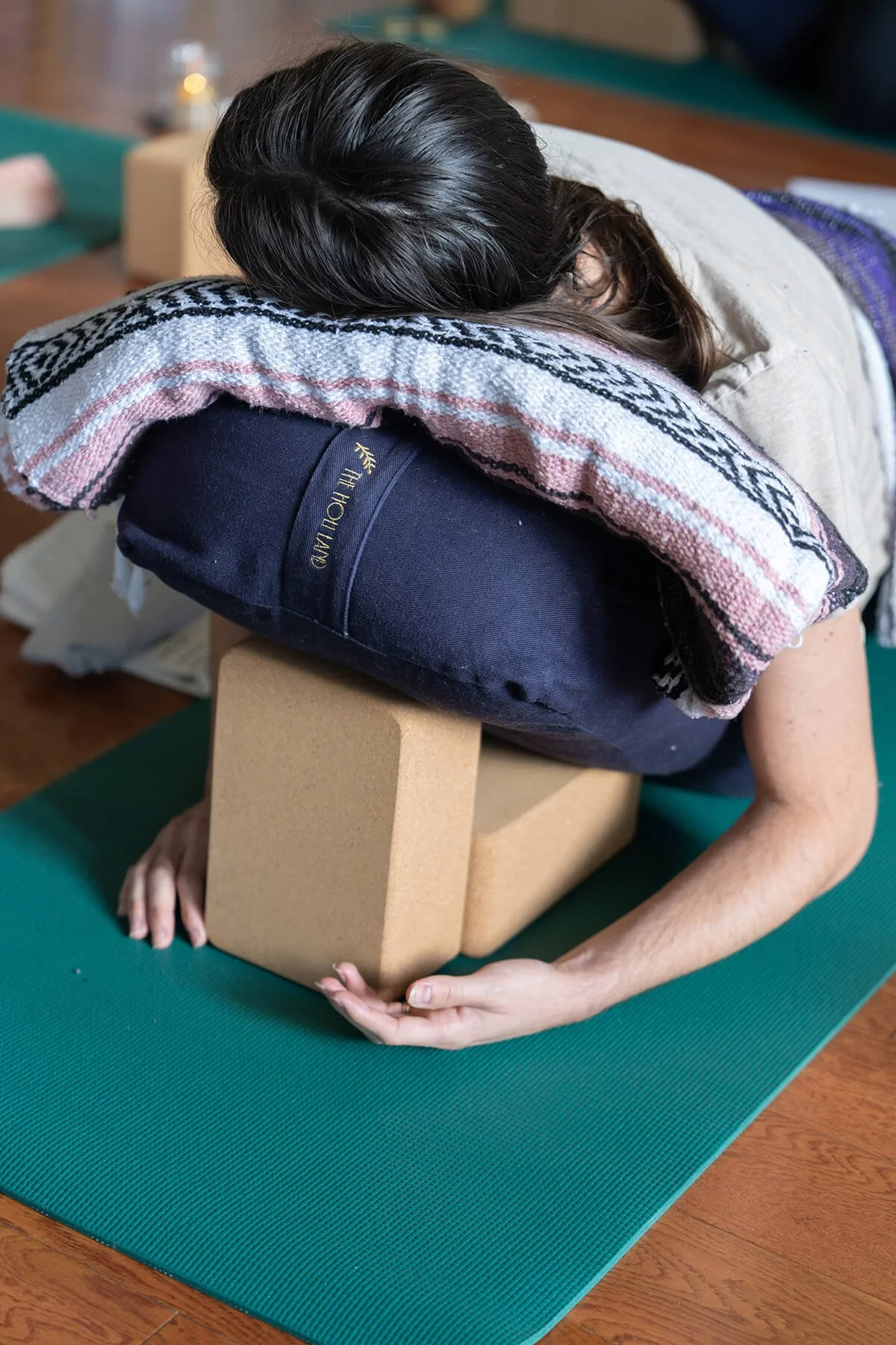 Person performing yoga pose on a green yoga mat, resting their head and arms on a cork yoga block with a towel on their back, in a room with wooden floors.