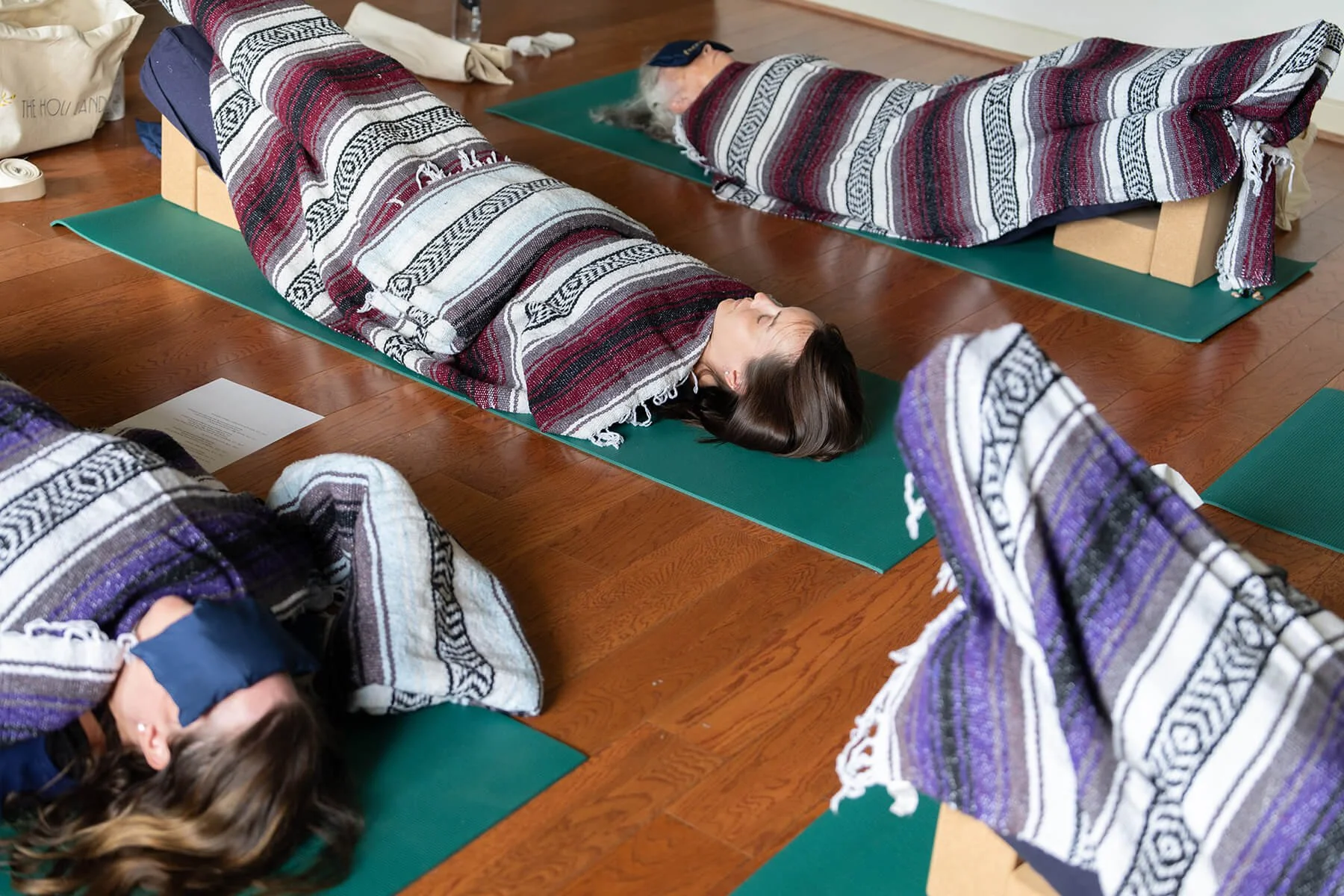 People lying on yoga mats, covered with blankets, participating in a relaxation or meditation session indoors on a wooden floor.