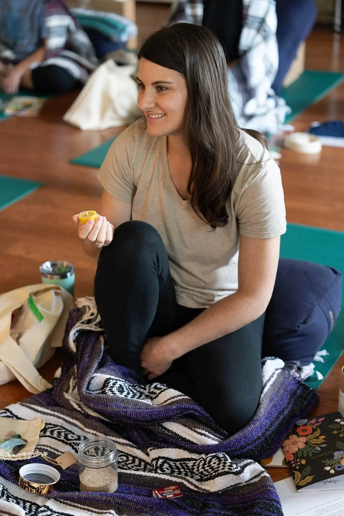 A woman with long dark hair sitting on a blanket indoors, smiling and holding a small container, surrounded by various items including jars, paper, and a book.