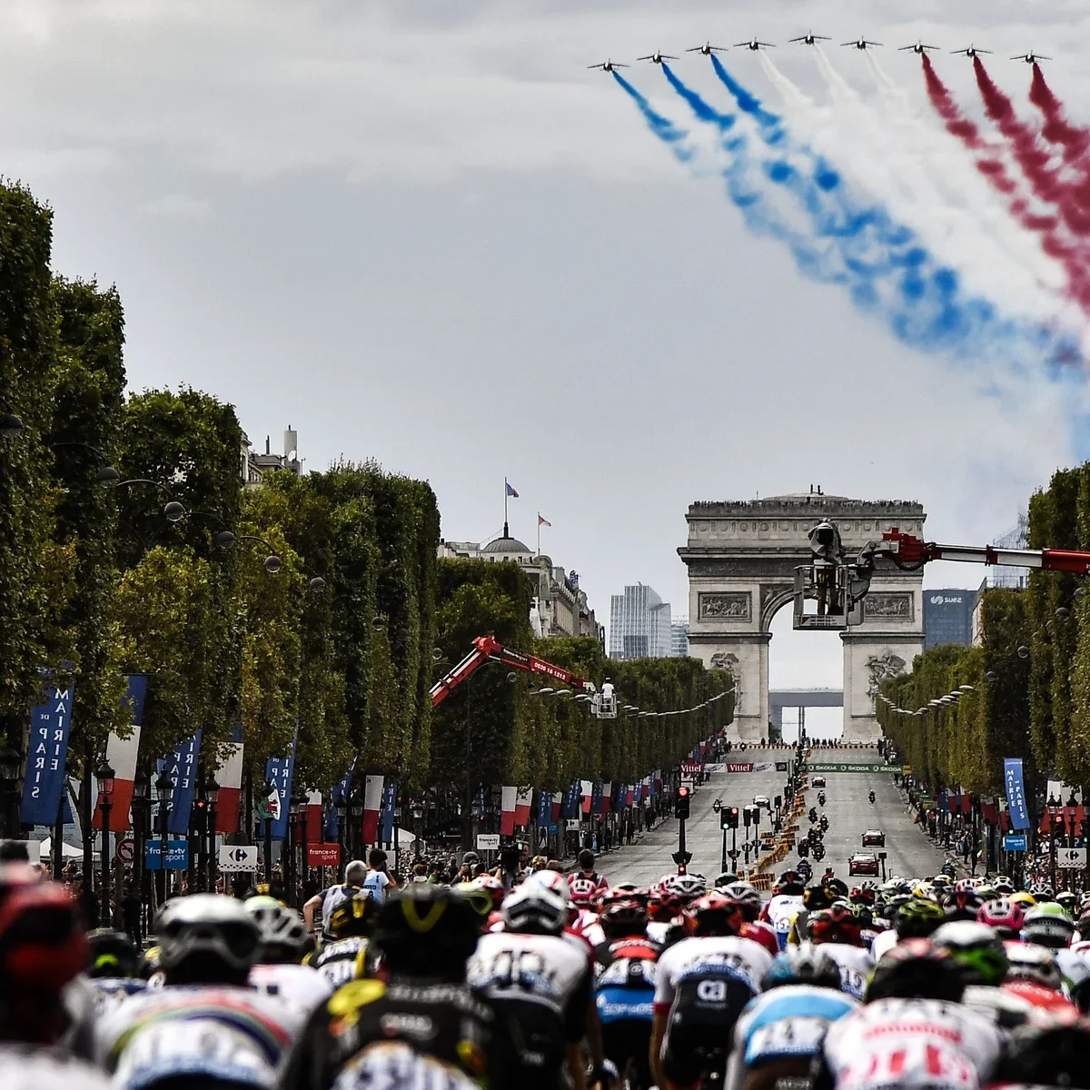 Un peloton de cyclistes dans une course sur l'avenue des Champs-Élysées avec l'Arc de Triomphe en arrière-plan, des avions civils volant dans le ciel en laissant des traînées de fumée tricolore (bleu, blanc, rouge) lors d'une célébration nationale.