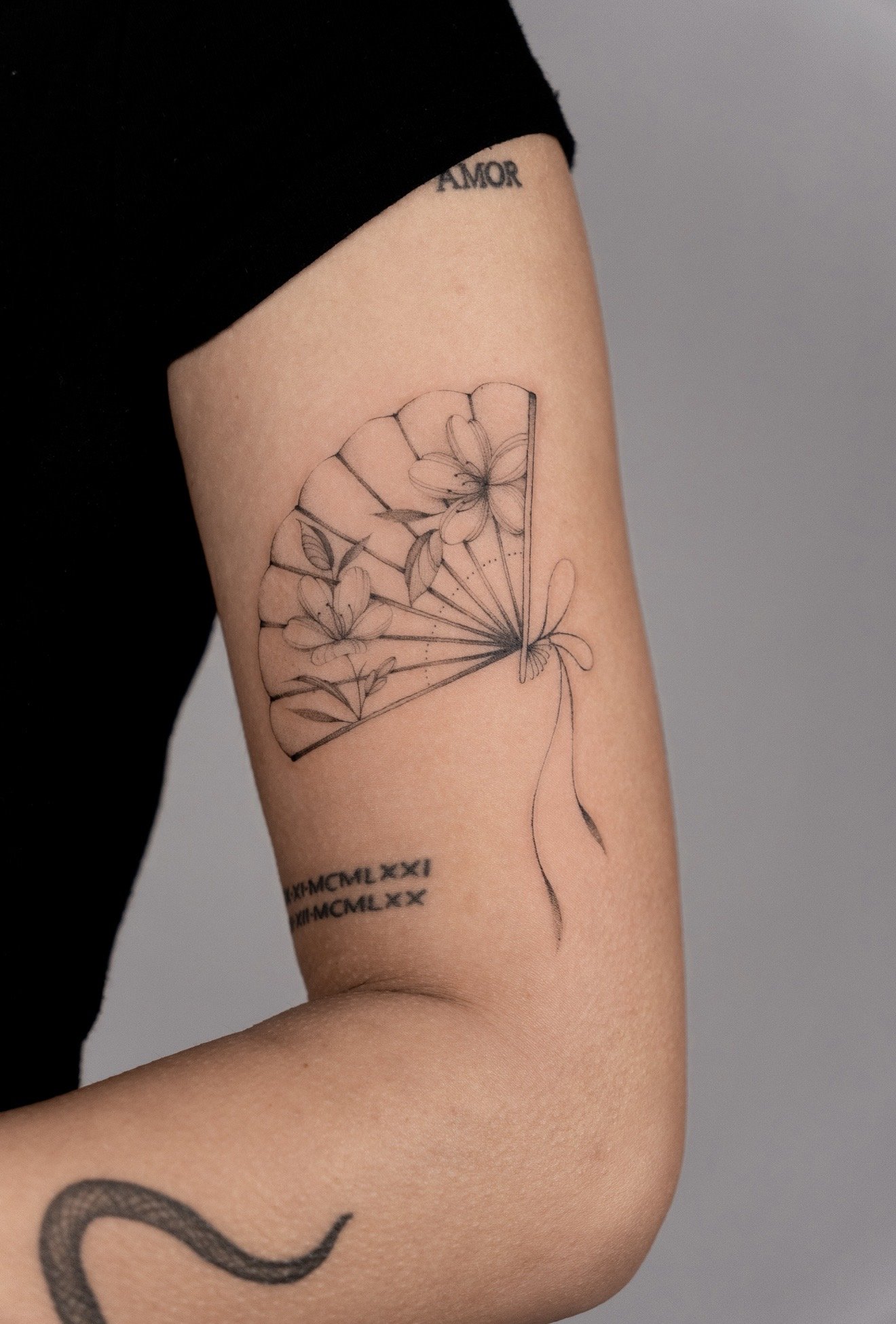 Close-up of a person's upper arm with tattoos, including a minimalistic line drawing of flowers, Roman numerals, and the word 'AMOR' on the shoulder.