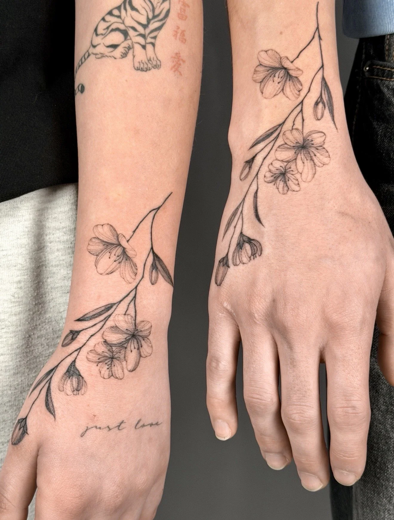 A close-up of two people holding hands, showcasing matching black and grey floral tattoos on their wrists and hands, with a dark background.