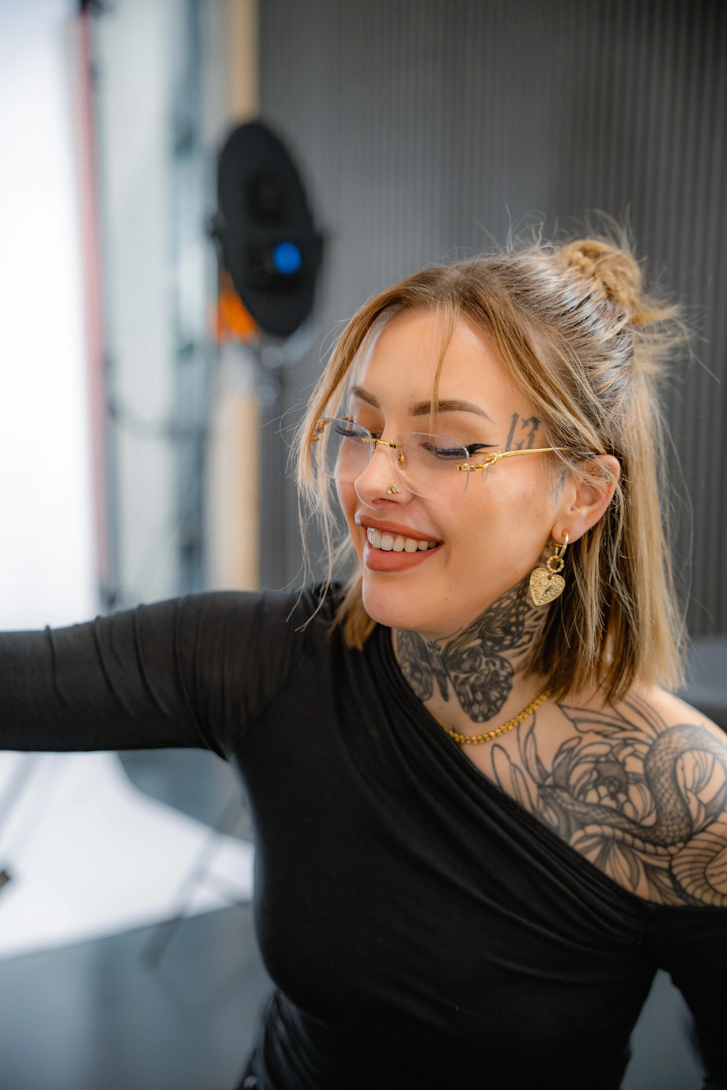 Close-up of a woman with light brown hair styled in a messy bun, wearing glasses with gold frames, earrings, and a nose ring, smiling while taking a selfie in a modern indoor setting.