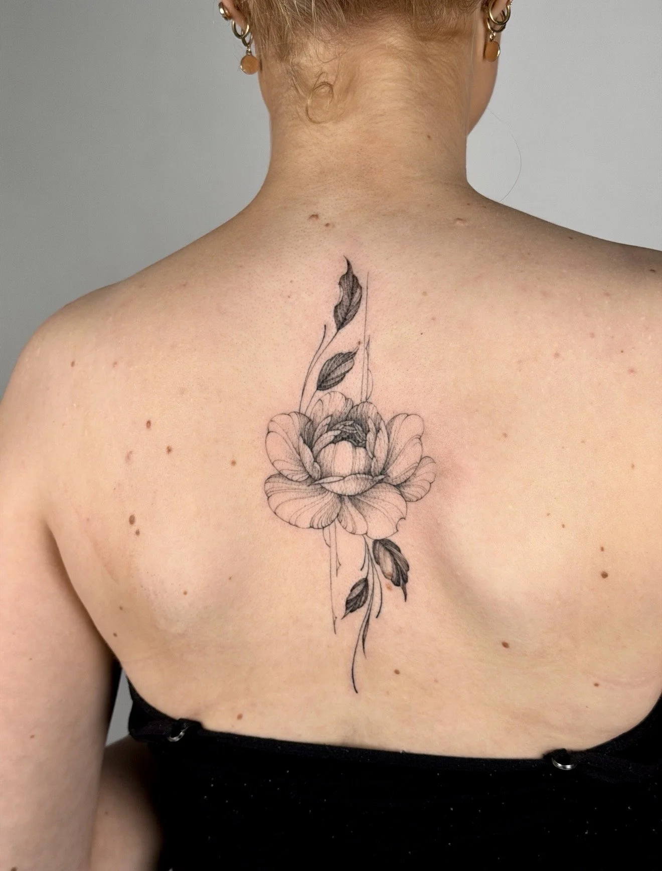 A woman's back with a black and grey floral tattoo featuring a blooming peony flower and leaves, located on the upper back/shoulder area.