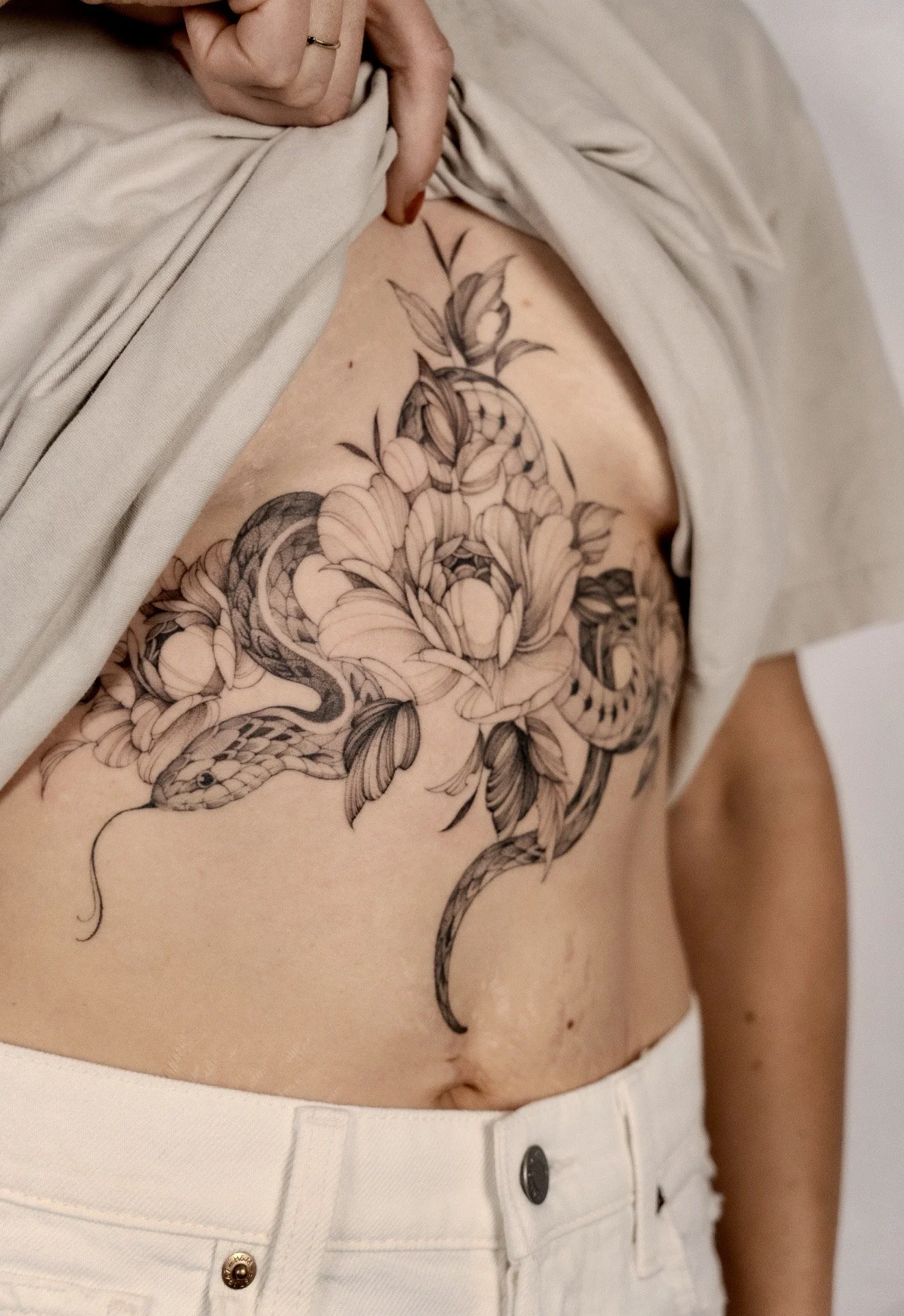 A person's torso with a detailed black and gray floral tattoo, partially covered by a beige shirt, and wearing high-waisted white pants.