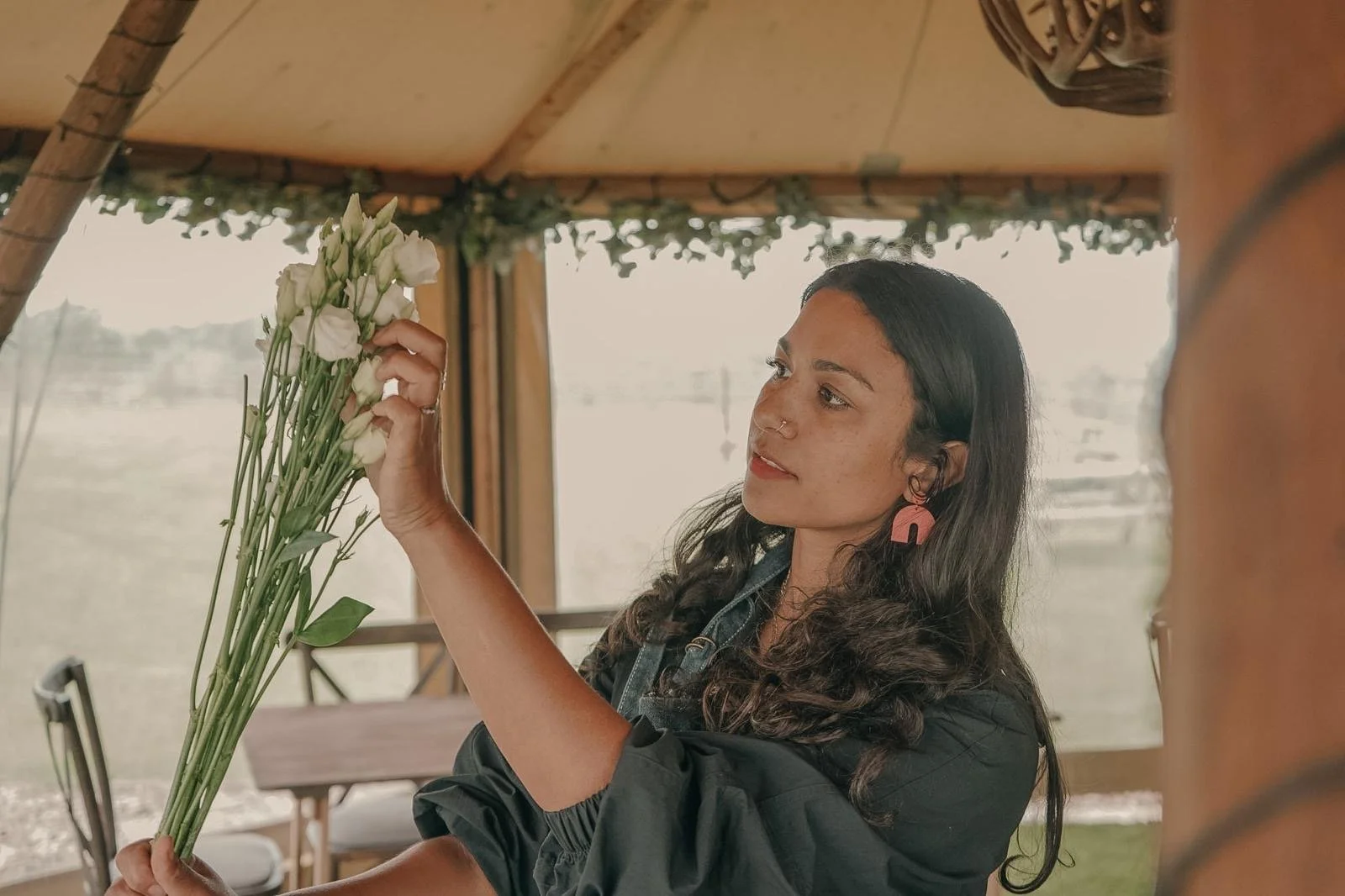 A woman with dark hair and earrings holding a bouquet of white and green flowers inside a wooden structure overlooking a body of water.