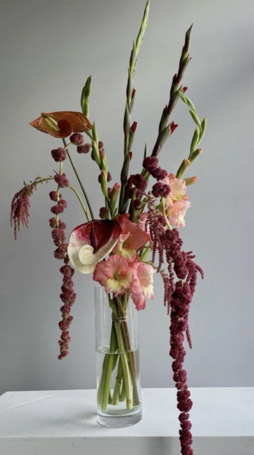 A tall glass vase holding a mixed floral arrangement with pink, red, and peach flowers, including lisianthus, calla lilies, and hanging amaranthus, set on a white surface against a plain gray background.