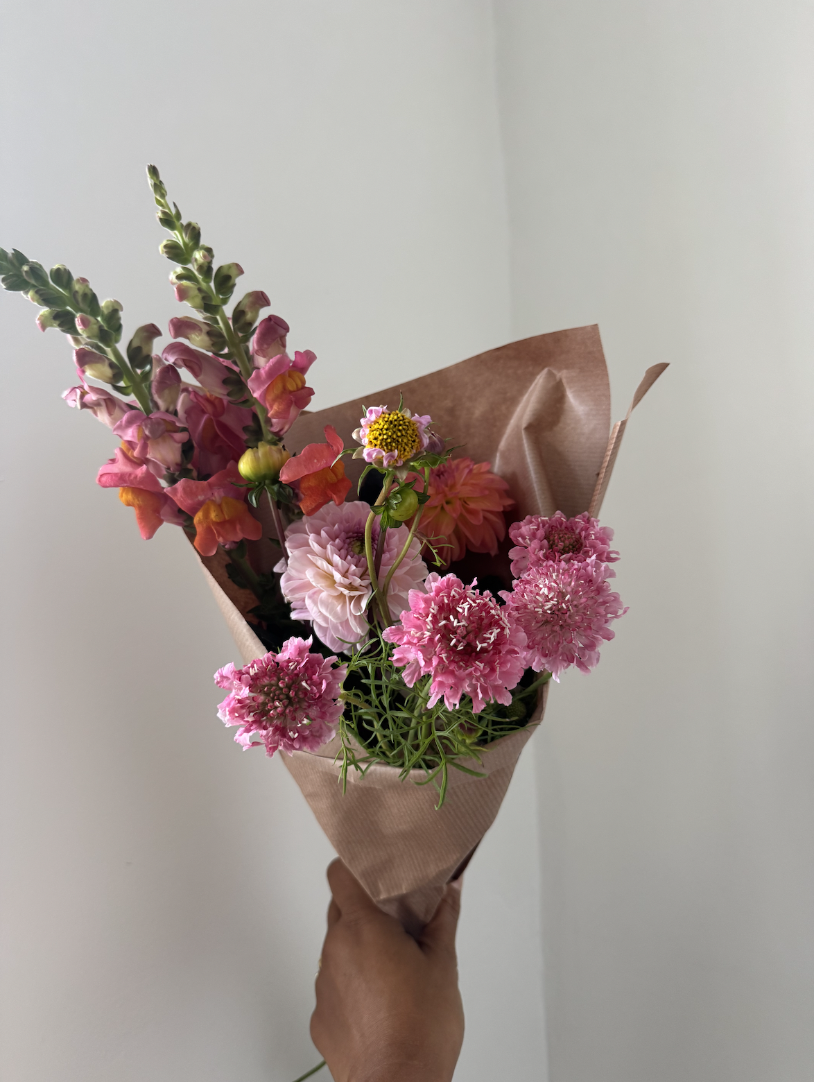 Hand holding a bouquet of pink, peach, and yellow flowers wrapped in brown paper against a plain white wall.
