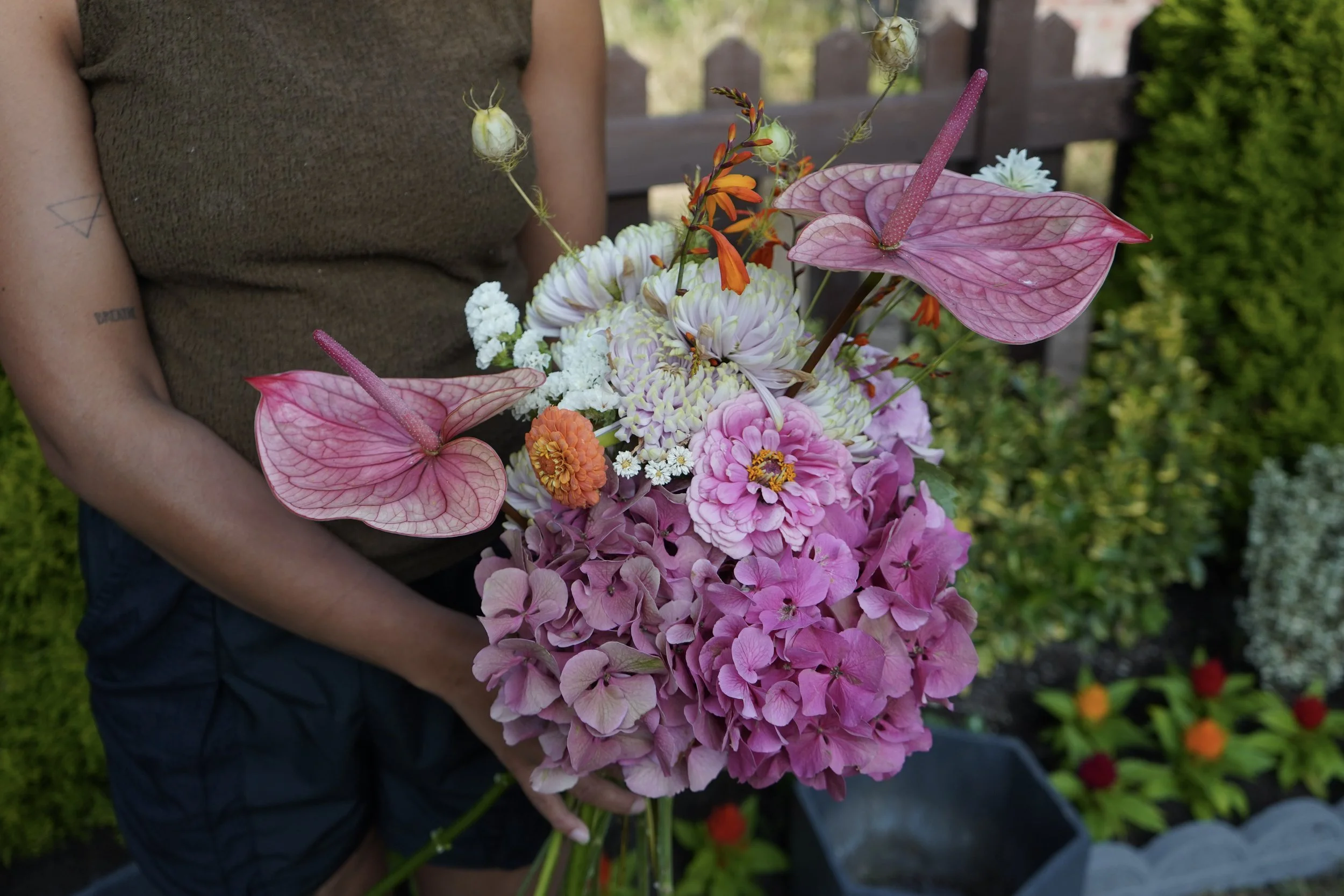 Person holding a colorful bouquet of flowers including pink anthuriums, white daisies, orange marigolds, and purple hydrangeas, with a blurred background of foliage and a wooden fence.