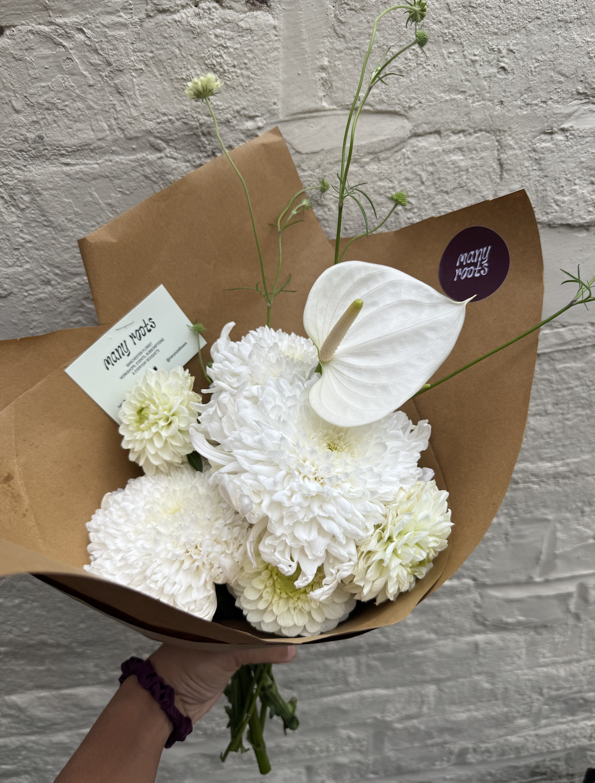 A bouquet of white flowers with a white anthurium, dahlias, chrysanthemums, surrounded by paper wrapping and a tag that reads "many roots," held against a textured white brick wall.