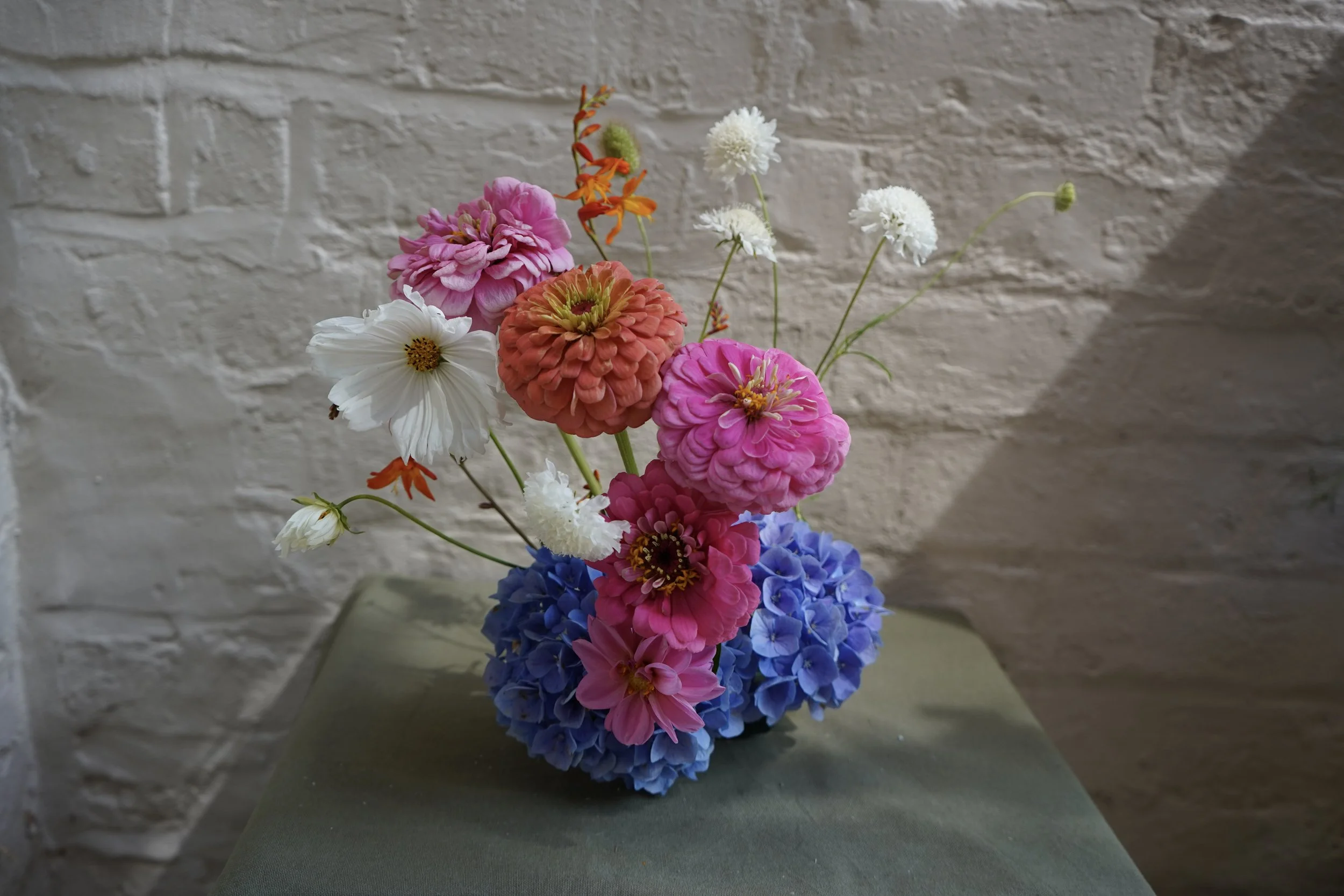 A bouquet of mixed colorful flowers, including pink, orange, white, and blue blooms, arranged in a vase against a white brick wall background.