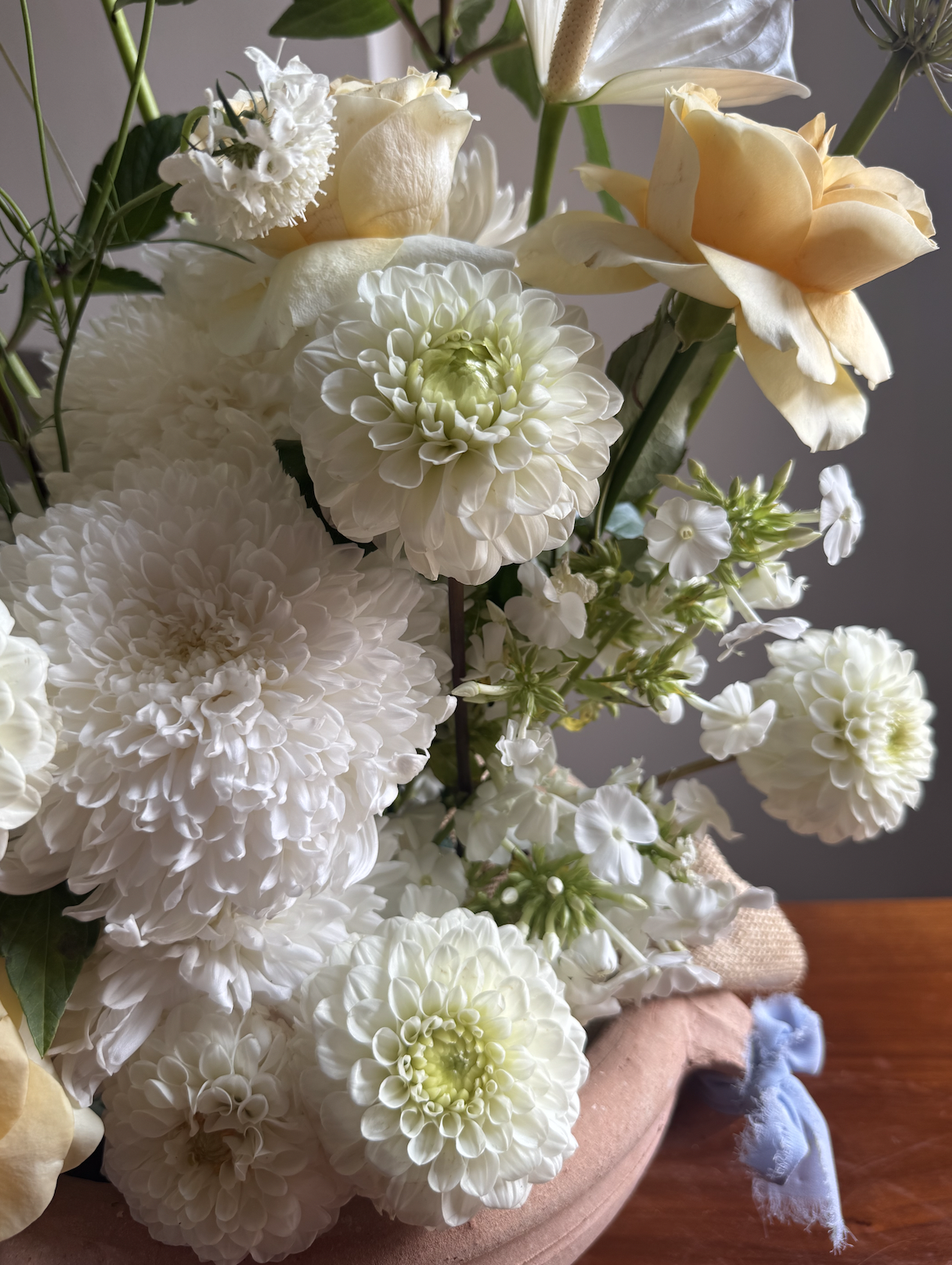 A close-up of a floral arrangement featuring white dahlias, cream-colored roses, and white lilies in a pinkish-orange vase, placed on a wooden surface.