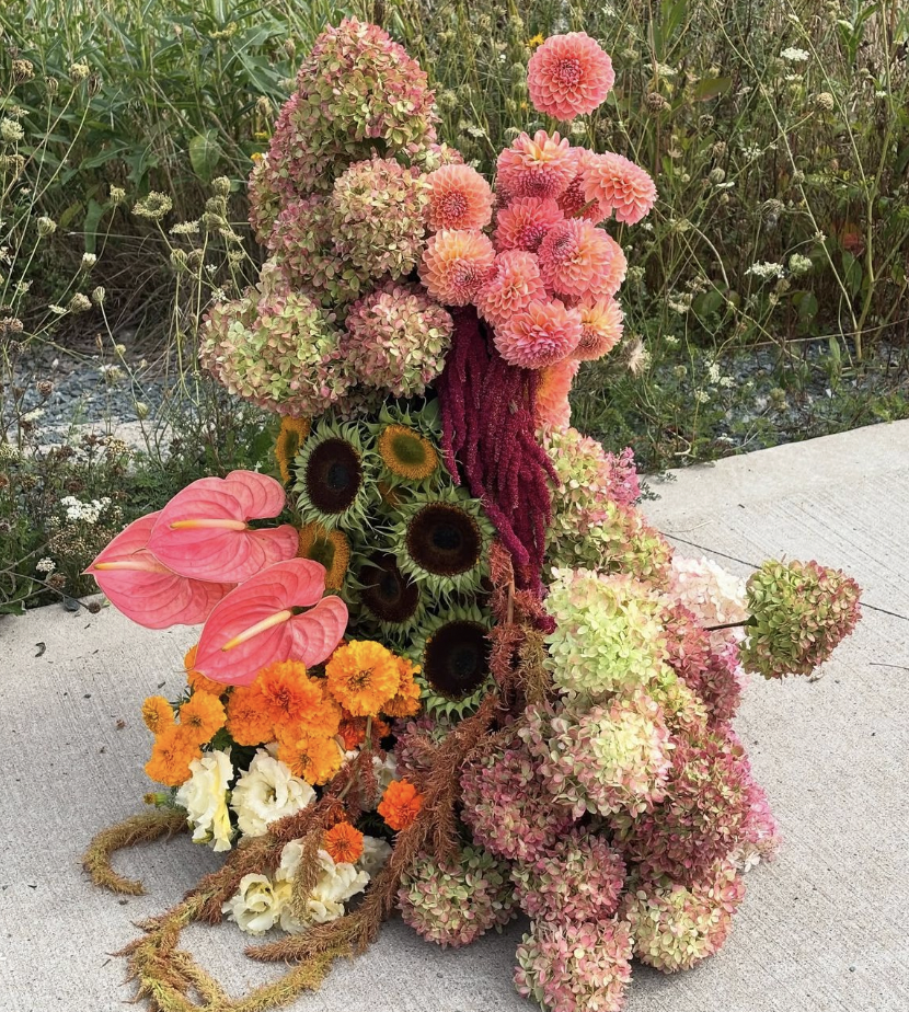 Colorful floral arrangement with hydrangeas, dahlias, sunflowers, anthuriums, marigolds, and amaranth, positioned on a concrete surface outdoors.