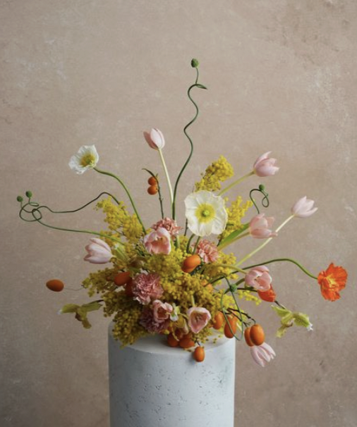 A colorful flower arrangement in a gray vase, featuring pink tulips, yellow flowers, white flowers, and orange buds, against a beige background.