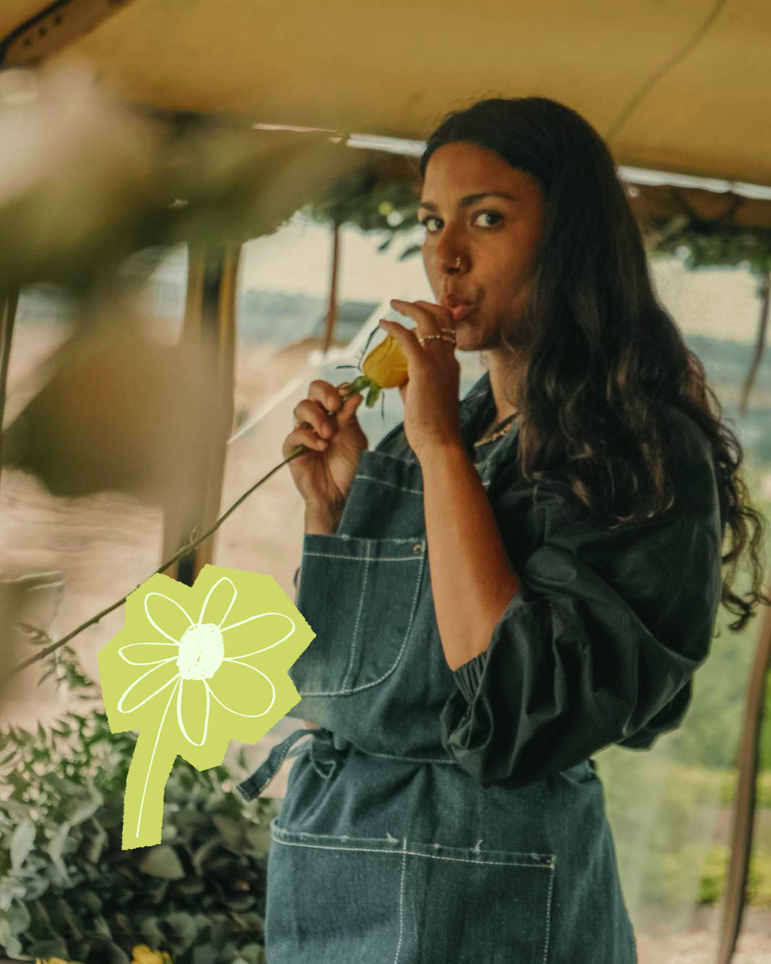 A woman with long dark hair, wearing a denim dress, is holding a yellow flower close to her lips and using a finger to shush, standing inside a greenhouse or garden space.
